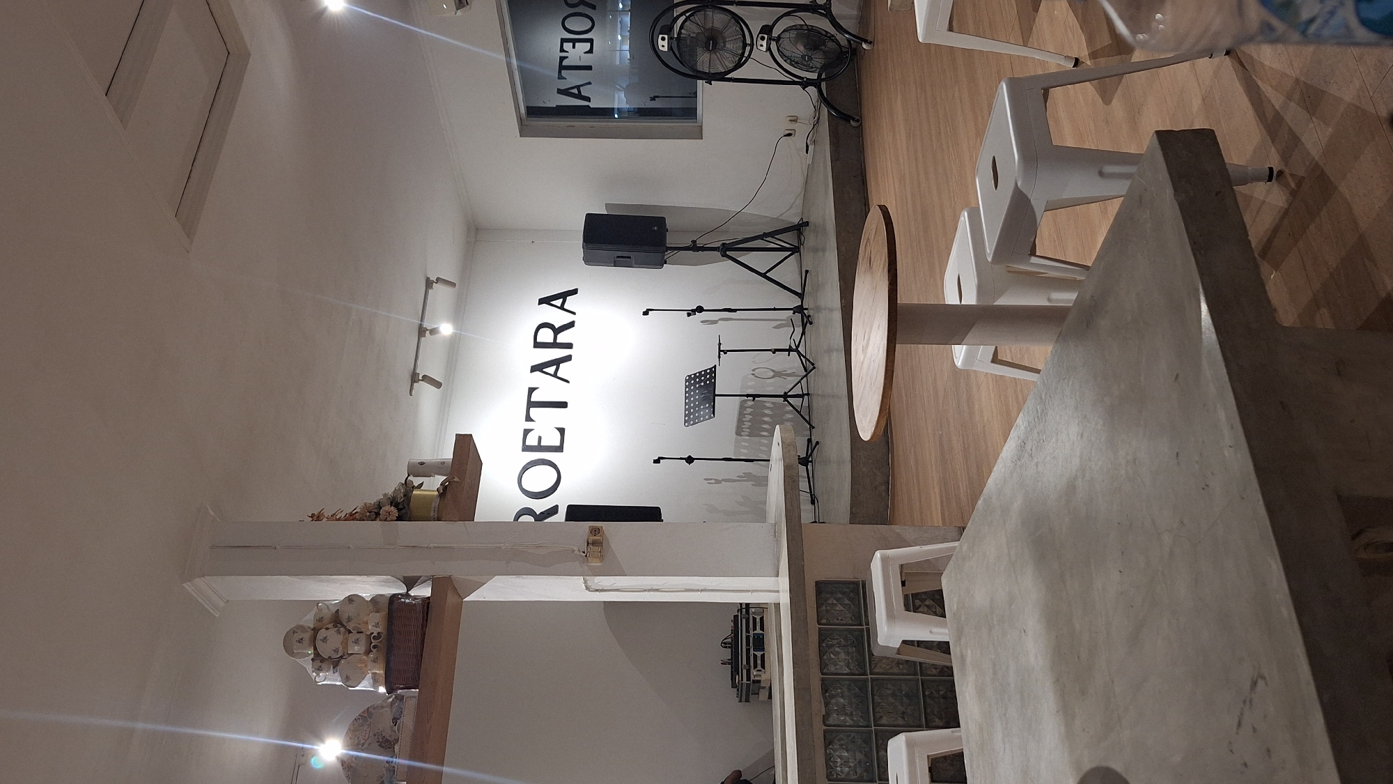 This interior shot features a stage with the word "ROETARA" vertically written on the wall. The setup includes a speaker, microphone stands, and a music stand. In the foreground, there's a wooden table with white chairs. A concrete table is also visible. The scene is likely located in North Jakarta, Indonesia, as suggested by the presence of "ROETARA", which may be a location-specific reference. There are no people explicitly depicted in the image.