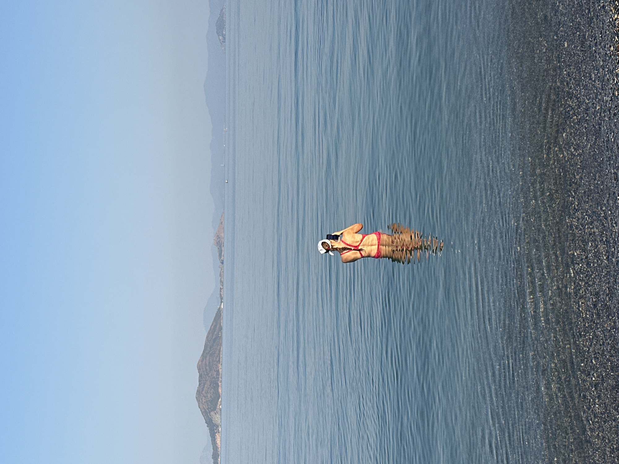 A woman stands in the ocean in Fethiye, Turkey, holding a smartphone to take a picture. She is wearing a red bikini and a white cap. The water is calm with gentle ripples. In the background, there are islands and a clear blue sky. The seabed is visible through the clear water, covered in small, dark pebbles.