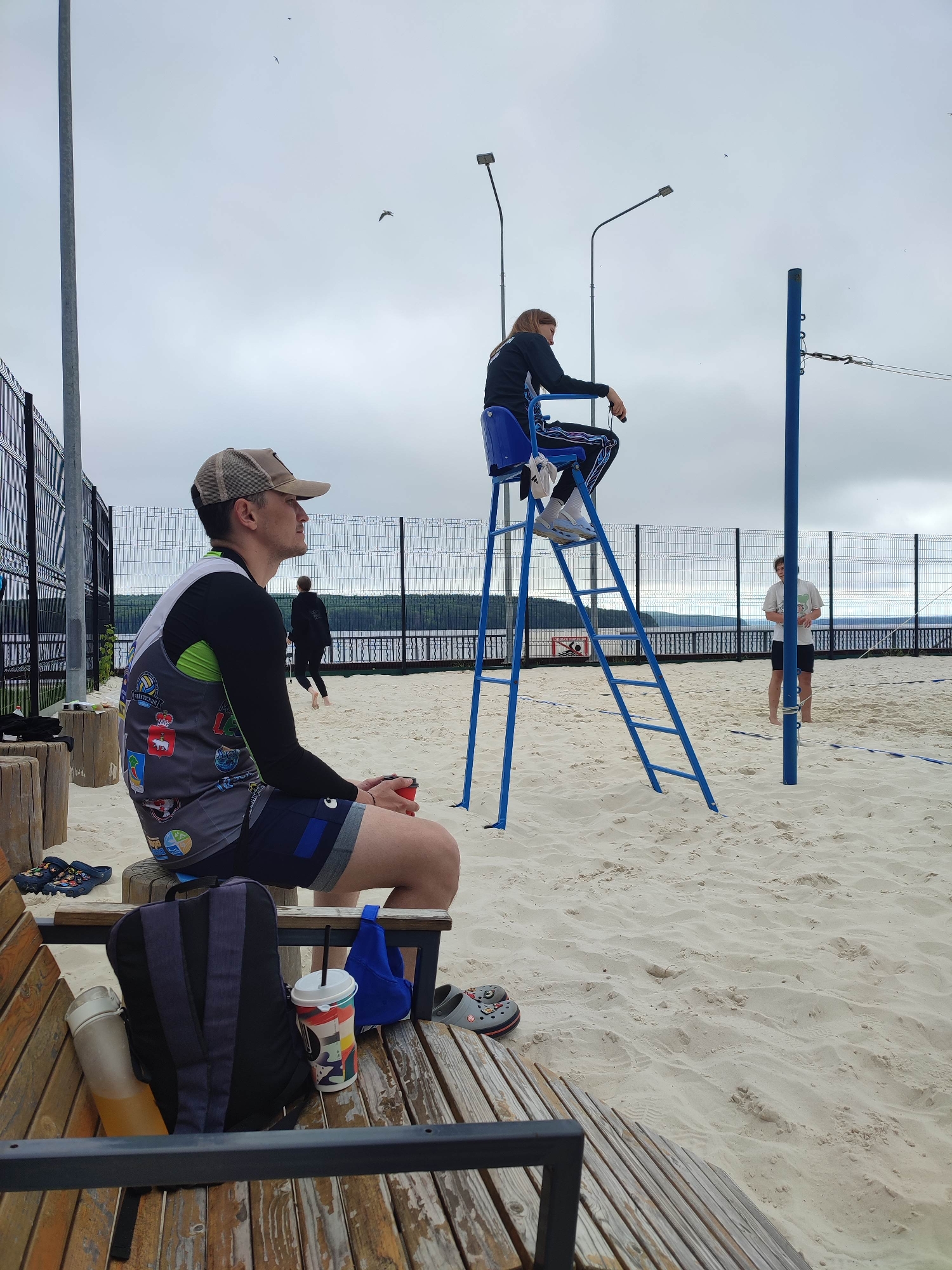 A volleyball referee sits on a high chair overlooking a sandy court. The referee, wearing a black jacket and pants, looks down at their phone. In the foreground, a man in a sleeveless shirt and cap sits on a wooden bench, holding a red cup. A backpack and drinks are placed on the bench next to him. Other people are visible on the court, including a person in the background running. The scene is set on a cloudy day with a body of water and trees visible in the distance.