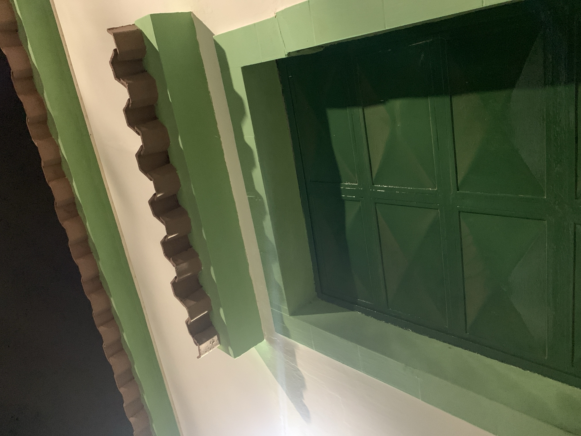 The image shows a section of a building exterior, possibly in Santa Ana, Venezuela, though this cannot be confirmed from visual information alone.
A green-framed window with a multi-pane grid pattern dominates the right side of the frame. The window is closed and heavily shadowed.
To the left of the window, the wall is white. Trim in two shades of green runs diagonally across the image, suggesting the building's roofline or architectural details. The inner trim is a darker, scalloped design. The darker area to the left is likely night sky.