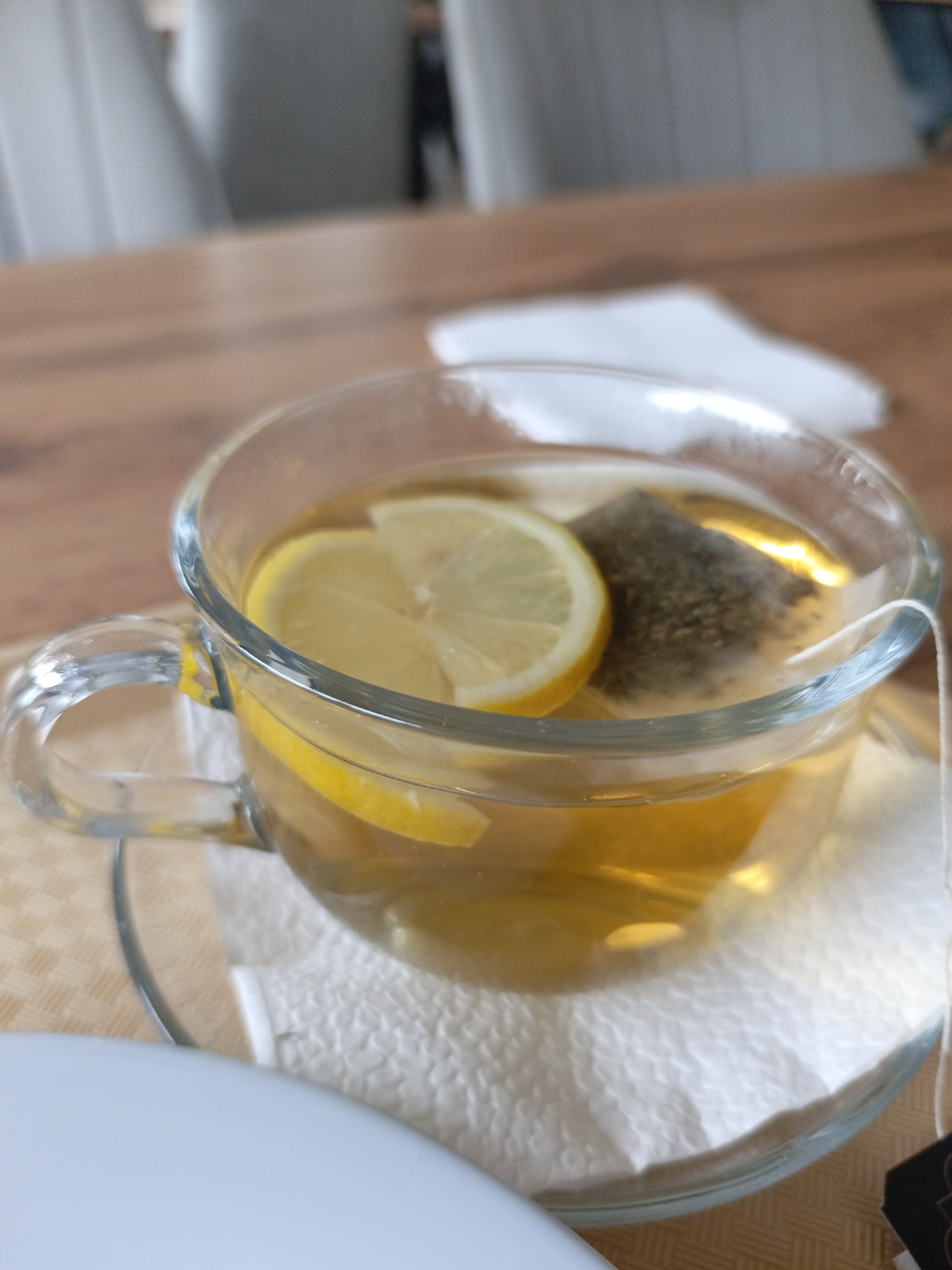 The specific location within Noviy, Russia, cannot be ascertained from the visual data presented. The scene is set indoors, indicative of a commercial dining establishment or private residence, with a light-brown wooden table as the primary surface.
The central subject is a clear glass mug, half-filled with an amber-colored liquid, presumed to be hot tea. Floating on the liquid surface are two circular slices of yellow lemon. A pyramid-shaped tea bag, containing coarse, dark green or brown botanical matter, is partially submerged in the liquid, with its white string extending over the mug's rim. The mug rests on a clear glass saucer, which in turn is placed on a white, textured paper napkin. Beneath the napkin and saucer, a light-brown woven placemat pattern is visible on the wooden table surface. In the immediate foreground, a partial white, curved object, likely a ceramic plate, is visible at the bottom left. The background is out of focus, revealing blurred forms of light-colored, high-backed chairs and indistinct structural elements. No individuals are visible.