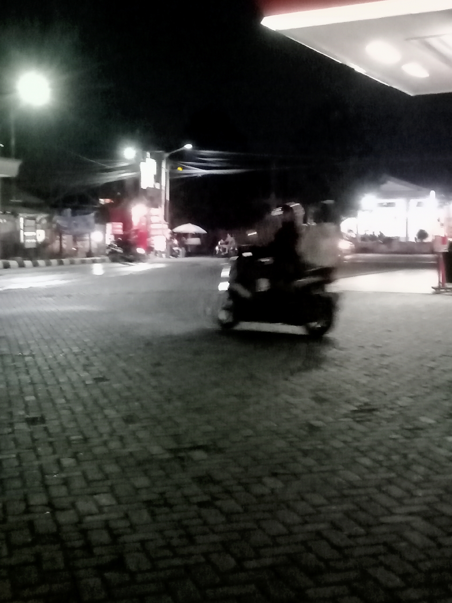A nighttime outdoor scene in Plosokandang, Indonesia, shows a paved area with brickwork, likely a street or public space. A single motorcycle, with a rider and passenger, is the main focus, captured in motion with motion blur. The motorcycle appears to be a scooter. Behind the motorcycle, the blurred lights of buildings and possibly a gas station canopy suggest an urban or semi-urban environment. A bright street lamp is visible on the left side of the frame, illuminating the scene. The overall atmosphere is dark, with artificial lighting creating highlights and shadows.