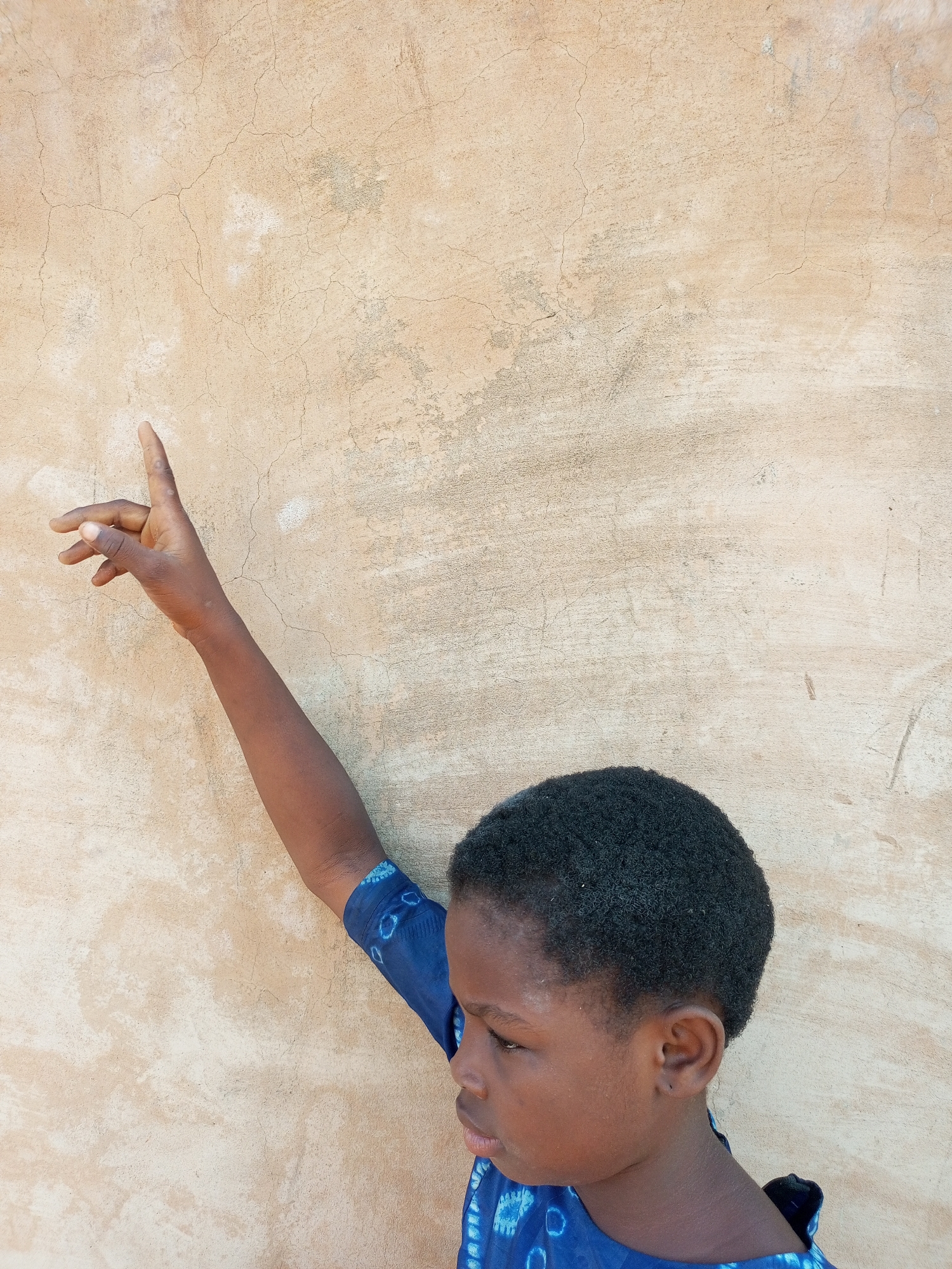 A young Black person is shown from the chest up, standing with their back to a textured, tan wall. The person is wearing a blue garment with a white pattern. Their right arm is extended upwards and to the left, with their index finger pointing. Their head is turned to the right, and their gaze is directed downwards. The background is a wall with visible cracks and variations in color.