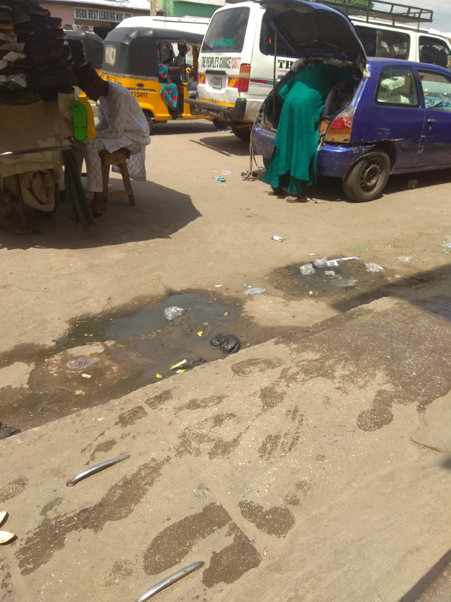 The image depicts a busy street scene in Jalingo, Nigeria, likely during daylight. In the foreground, a puddle of dirty water with some scattered trash, including plastic bags and wrappers, reflects the harsh sunlight. The ground is a mix of concrete and dirt, with some subtle paving stones visible.
Several vehicles are present. To the left, a yellow auto-rickshaw, or "Keke Napep," with a license plate ending in "T541T," is parked. Next to it, a man in traditional white attire, including a cap, is seated on a low stool, appearing to rest or wait. He is positioned near a stall filled with dark items, possibly merchandise like bags or clothing, partially obscured by a large shadow.
In the center and to the right, a blue hatchback car is parked with its trunk open. A person wearing a long, teal-colored garment is bent over, rummaging through the trunk. Behind the car, a white passenger van with roof racks is visible. Text on the van reads "THE PEOPLE'S CHOICE PART". A smaller sign on a building in the background says "USBA BEST LEADING NIG".
The scene suggests a commercial or transportation hub. The presence of trash and the puddle implies an informal setting, possibly a market area or a roadside stop. The weather appears sunny, indicated by the strong shadows. The overall atmosphere seems active and bustling, typical of a Nigerian urban environment.