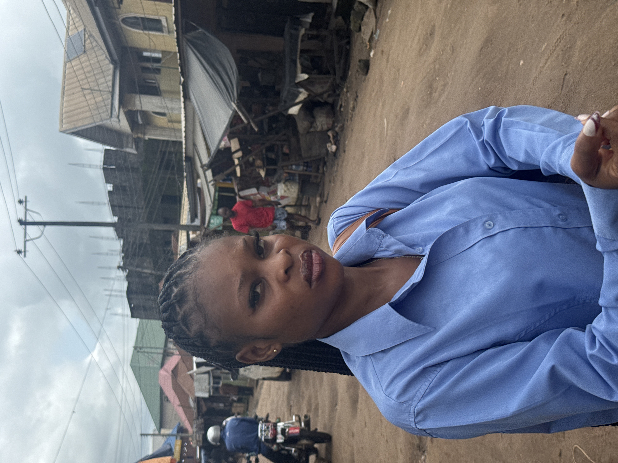 A young Black woman with braided hair and dark makeup poses for a close-up portrait in what appears to be a street market in Ihiagwa, Nigeria. She is wearing a light blue collared shirt and a brown strap is visible over her shoulder. Her expression is neutral. In the background, other people are visible, including a man in a red shirt walking away from the camera, and a person on a motorcycle. The environment is dusty with various market stalls and buildings. The sky is overcast.