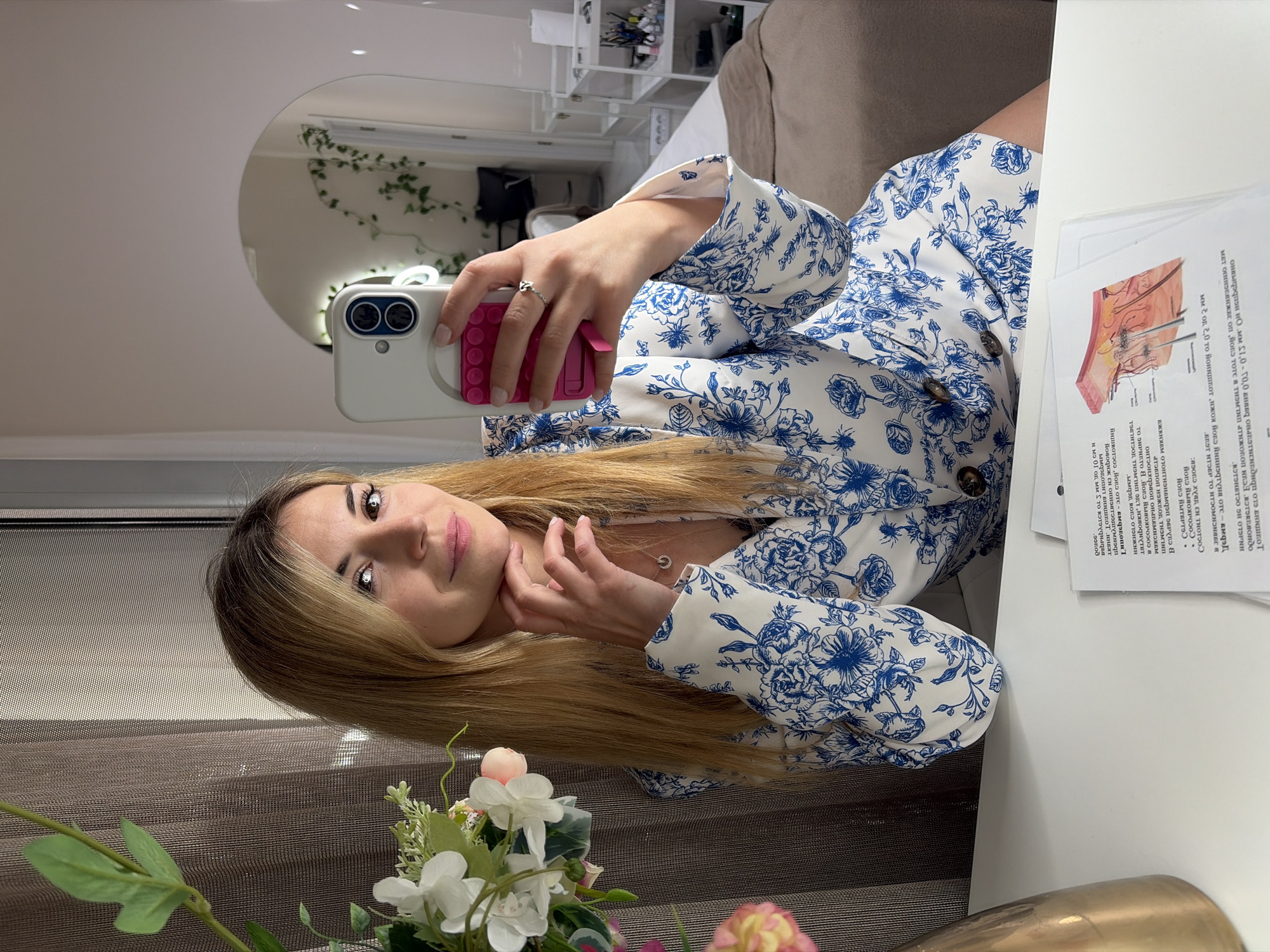 A woman is taking a selfie in an indoor setting. She is holding a white smartphone with a pink protective case, and her ringed finger is visible. She is wearing a white blazer with a blue floral pattern, a necklace, and her blonde hair is cascading to the side. In the background, there's a mirror with ivy, and shelves with various items. There's also a table with documents and a vase with flowers. The setting appears to be in Budva, Montenegro, potentially a salon or a similar establishment offering beauty treatments, based on the documents with diagrams and text in a Cyrillic alphabet. The woman seems to be posing for the photo.