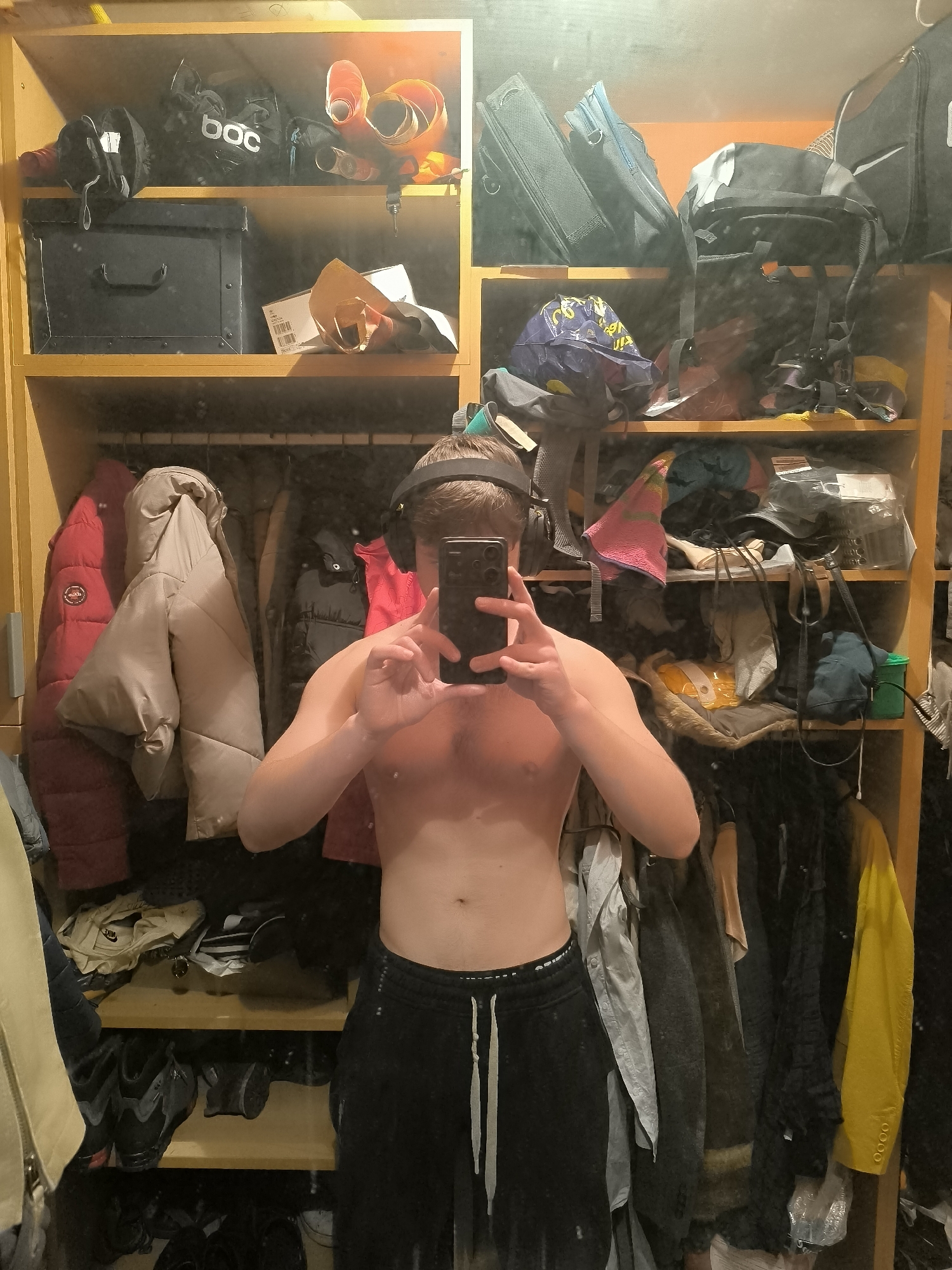 A person is taking a shirtless selfie in a mirror, revealing a cluttered closet. The closet is filled with shelves containing luggage, backpacks, skiing gear, and boxes. There are also clothes hanging on a rod, including jackets and coats. The person is wearing headphones and black sweatpants with white drawstrings. The image provides no information to determine the city of Sofia, Bulgaria, as the location.