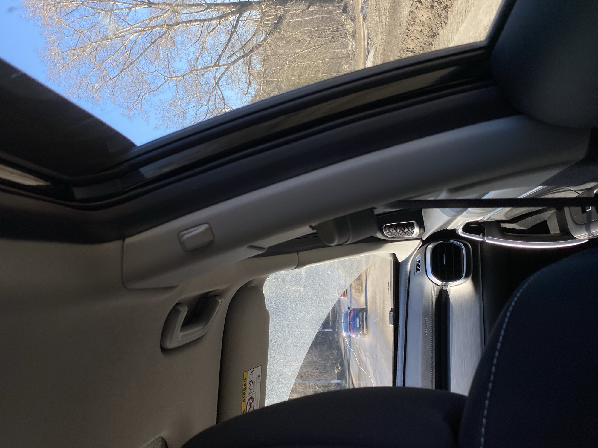 The image shows the interior of a car with a sunroof. The sunroof is open, revealing bare tree branches against a bright blue sky. A car seat and a portion of the dashboard are visible in the foreground. Looking out of the side window, one can see a road with another vehicle on it. Based on the presence of a sun visor with warning labels, and the style of the car interior, it is likely a modern passenger vehicle. The image does not provide enough information to confirm the specific location as Chaikovsky, Russia, or any specific location, or details of people, subjects, objects, actions, or interactions not already described.