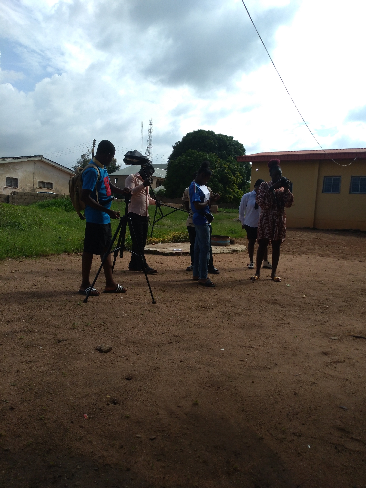The image captures an outdoor scene in Jos, Nigeria, showing five individuals engaged with camera equipment on reddish-brown dirt ground, set against a backdrop of buildings and vegetation under a heavily overcast sky.
The ground in the foreground is dry, uneven earth with scattered small stones and faint impressions. Patches of green grass are visible to the left of the central group of people and surrounding a partially obscured blue metal drum located in the mid-ground.
From left to right, the individuals are: A dark-skinned male wearing a blue t-shirt, black shorts, black-and-white striped slide sandals, and a beige backpack. He is positioned over a black tripod, actively adjusting its components with his hands. To his right and slightly behind, a second dark-skinned male, dressed in a light peach-colored long-sleeved shirt and dark trousers, also in dark slide sandals, holds a piece of black equipment, possibly a reflector or light diffuser, near the tripod. Partially obscured between these two males and further back, a third individual in blue trousers or jeans is visible, looking down at an object held in their hands. To the right of the tripod setup, a fourth dark-skinned individual, wearing a white long-sleeved shirt and dark shorts, stands with hands clasped, observing. The rightmost individual is a dark-skinned female with reddish-brown hair, styled in short twists or dreadlocks, wearing a patterned knee-length dress in shades of brown, red, and white, and gold-colored sandals. She is holding a black DSLR camera to her right eye, appearing to operate it.
In the background on the left, a single-story, light-colored building with a dark roof and two small, dark-framed windows is visible, with power lines connecting to it. To the right of this building, two tall, lattice-structure communication towers ascend into the sky. Further right and behind the group, a single-story yellow building with a reddish-brown roof features two prominent blue-framed windows. A large, dark green leafy tree stands behind the yellow building, extending above the central figures. A single black electrical wire traverses diagonally downwards from the upper right portion of the frame.
The sky is entirely covered by expansive grey clouds, providing a diffused, soft illumination across the entire scene, consistent with daytime conditions.