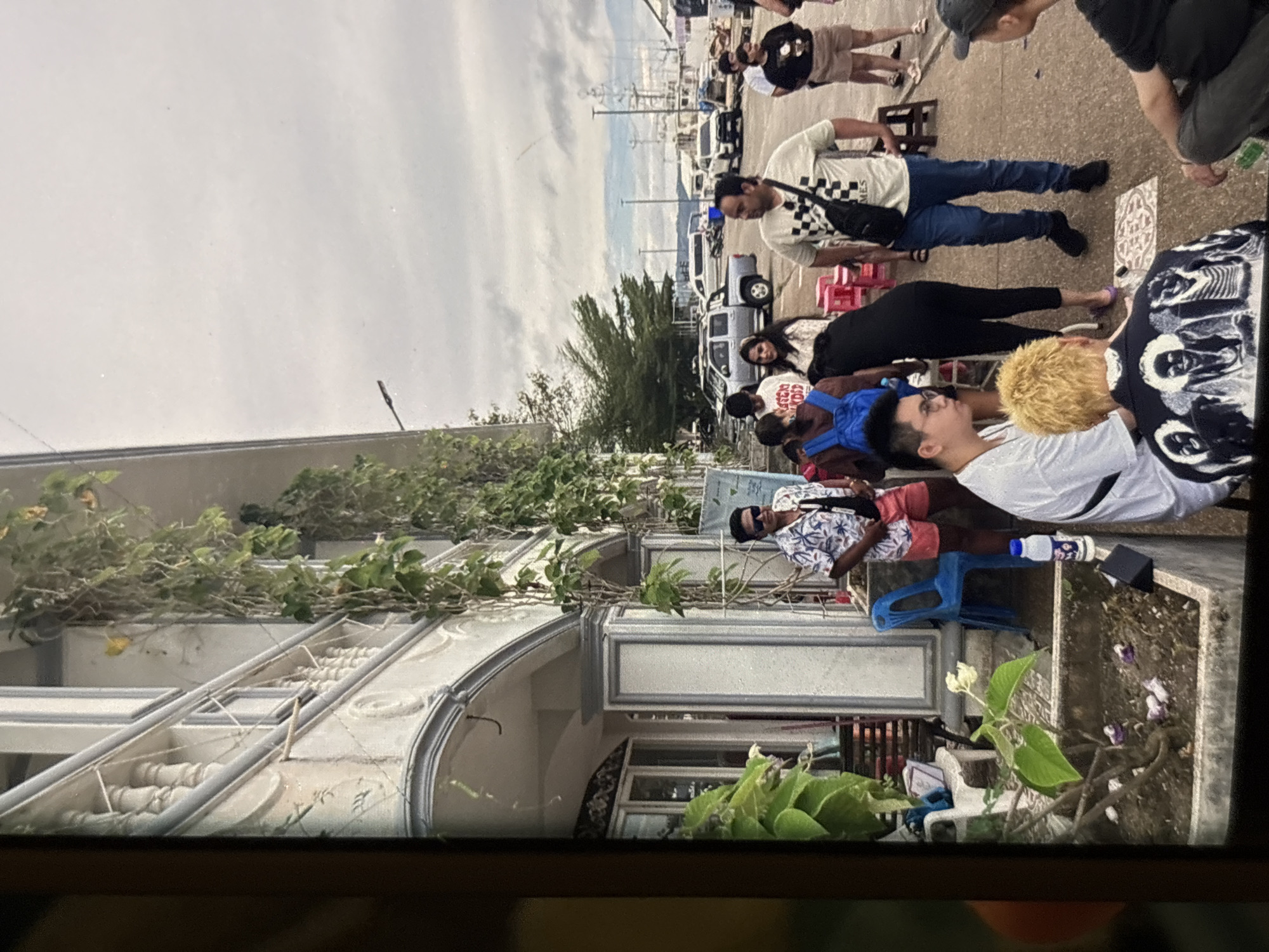 The image depicts a group of people gathered outdoors in Chalong, Thailand, with a building facade featuring elaborate architectural details and climbing greenery to the left. In the foreground, several individuals are standing and sitting. One man in the center wears a white t-shirt with a checkered pattern, blue jeans, and a black crossbody bag. To his right, a man in shorts and a patterned shirt stands. In the foreground, a person with bright blonde hair is seated, wearing a black jacket with a graphic print. A man wearing glasses and a white t-shirt is seated to the left. Behind them, other individuals are visible, some carrying bags. The background shows parked vehicles, including a truck and vans, and a body of water with boats, suggesting a harbor or pier area. The sky is overcast. The scene appears to be a public gathering or waiting area.