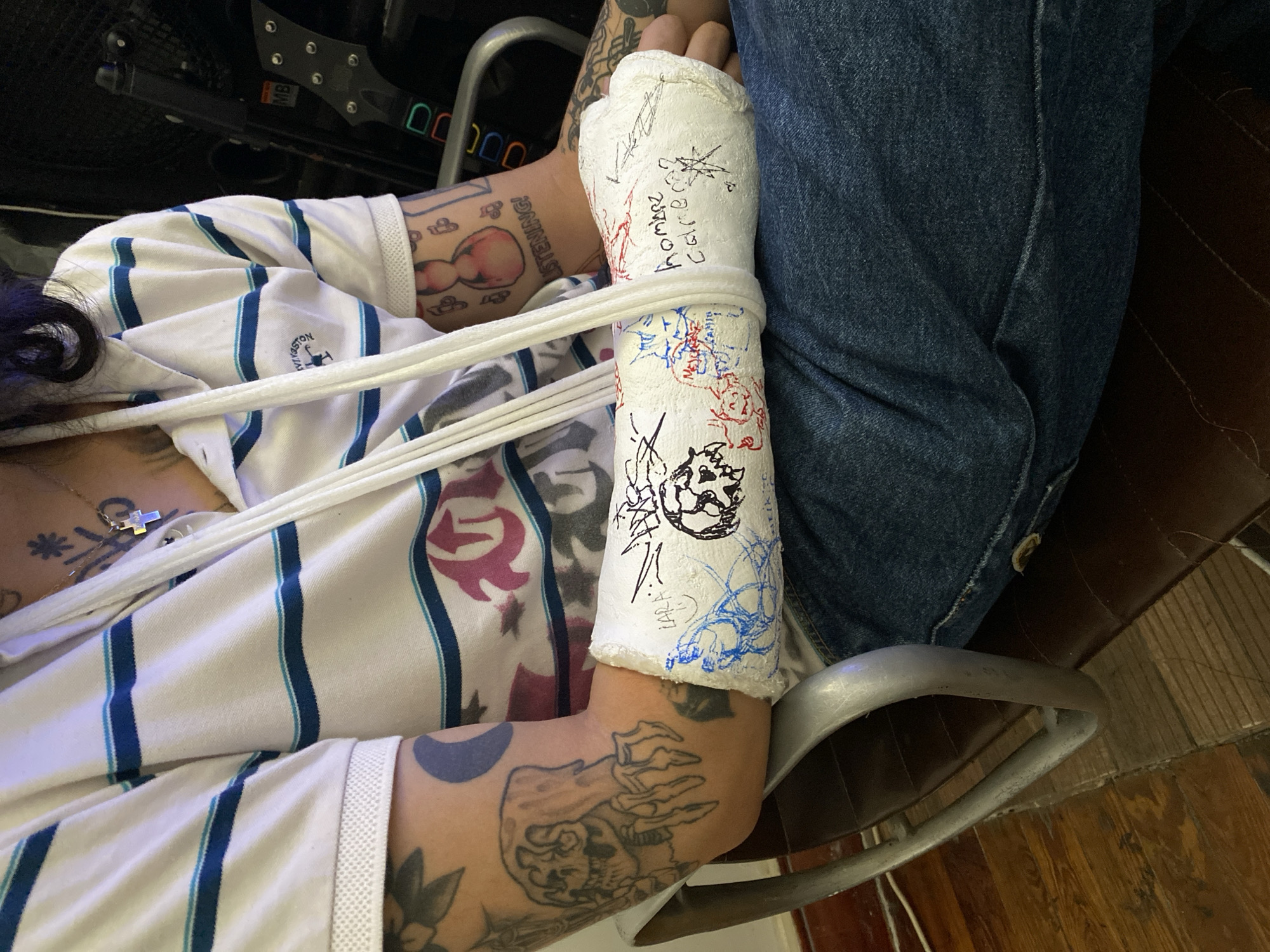 A person sits in a wheelchair, their left arm in a white cast. The cast is decorated with drawings in black, red, and blue ink, as well as signatures, including one that reads "Coloca". The person is wearing a white polo shirt with blue stripes and jeans. A white rope-like object is draped across their chest. Tattoos are visible on their arms and chest. The wheelchair has black and silver elements and sits on a hardwood floor. There is no clear evidence from the photograph to determine the location in Buenos Aires, Argentina.