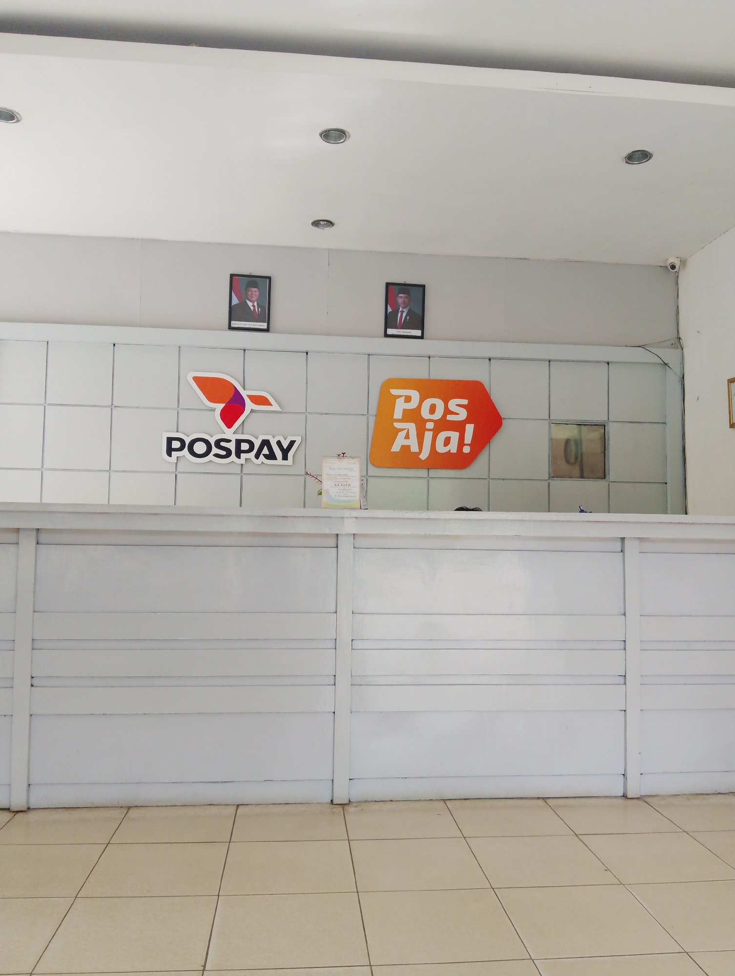 The image depicts the interior of what appears to be a Post Office or financial service counter in Serbelawan, Indonesia. The scene is dominated by a white reception desk with horizontal paneling. Behind the desk, a tiled wall features two prominent logos: one for "POSPAY" with an orange, purple, and white abstract bird-like graphic, and another for "Pos Aja!" displayed in white text on an orange arrow-shaped background. Above the logos, two framed portraits of Indonesian officials are mounted on the wall. A small security camera is visible in the upper right corner of the frame. A clear plastic display stand with some text is placed on the counter to the right of the POSPAY logo. The floor is covered with light-colored square tiles. The lighting appears to be from overhead fluorescent fixtures, and the overall environment suggests a public service space.