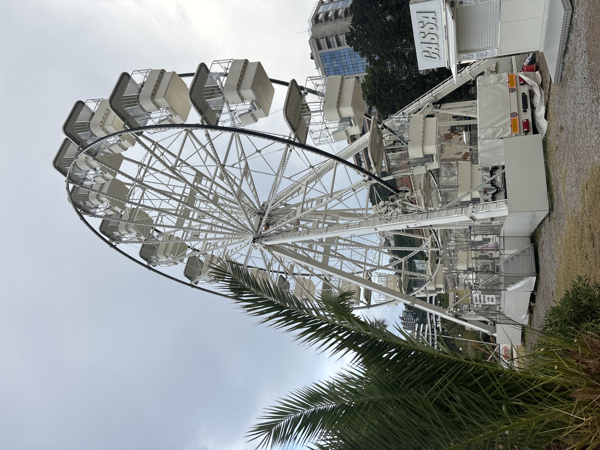 This media file depicts a large, white Ferris wheel situated outdoors. The Ferris wheel is the primary object in the scene, with its intricate metal framework and numerous passenger cabins visible. In the background, a modern building with glass windows and trees can be seen, suggesting an urban or semi-urban setting. The foreground is partially obscured by the fronds of a palm tree, adding a touch of nature to the scene.
The Ferris wheel appears to be stationary, with no visible activity of people riding it. The sky is overcast with a light gray color, indicating a cloudy day. The overall mood is neutral, with no strong emotions conveyed. The location cues, such as the palm tree and the style of the building, combined with the provided context of Budva, Montenegro, place the scene in that coastal city.
One piece of visible text is "CASSA" written in large white letters on a white sign, likely indicating a ticket booth or an entrance to the attraction. There might be other smaller signs, but they are not clearly legible. The time of day is indeterminate due to the overcast sky, but it appears to be daytime. The Ferris wheel is likely part of a temporary amusement setup, given its mobile appearance with wheeled support structures visible.