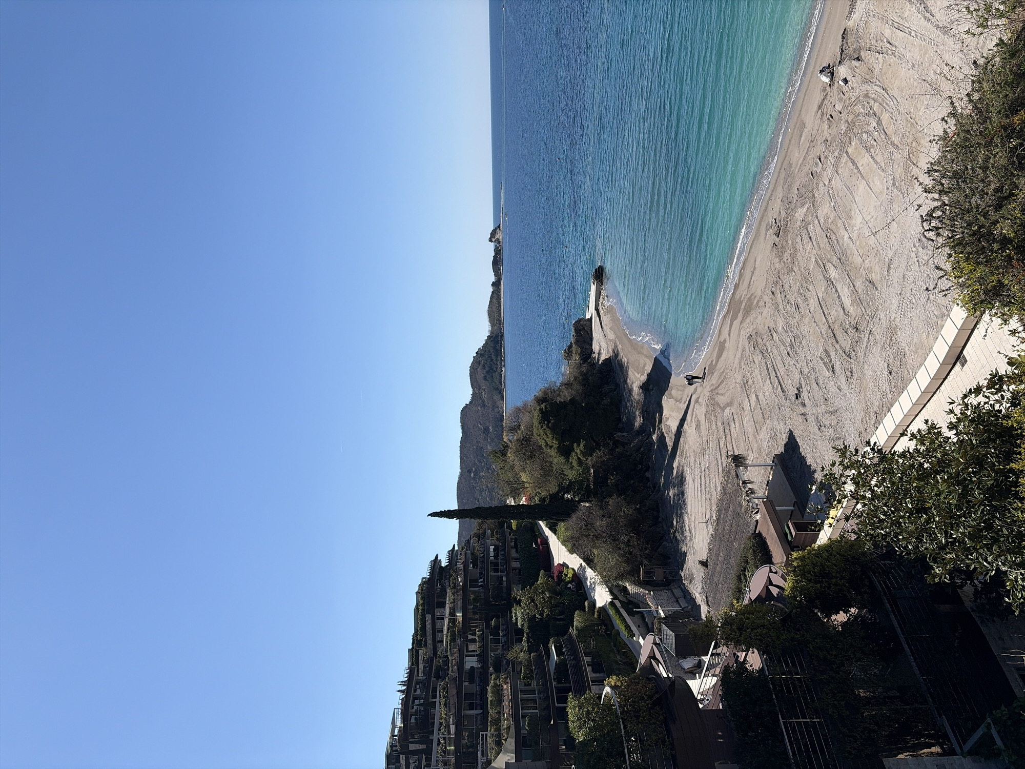 The image depicts a coastal scene featuring a beach with tire tracks in the sand, adjacent to a blue sea. A man is walking on a pier. On a slope above the beach is a densely built residential complex with vegetation. The sky is clear and blue. The location is in the city of Unknown, Unknown.