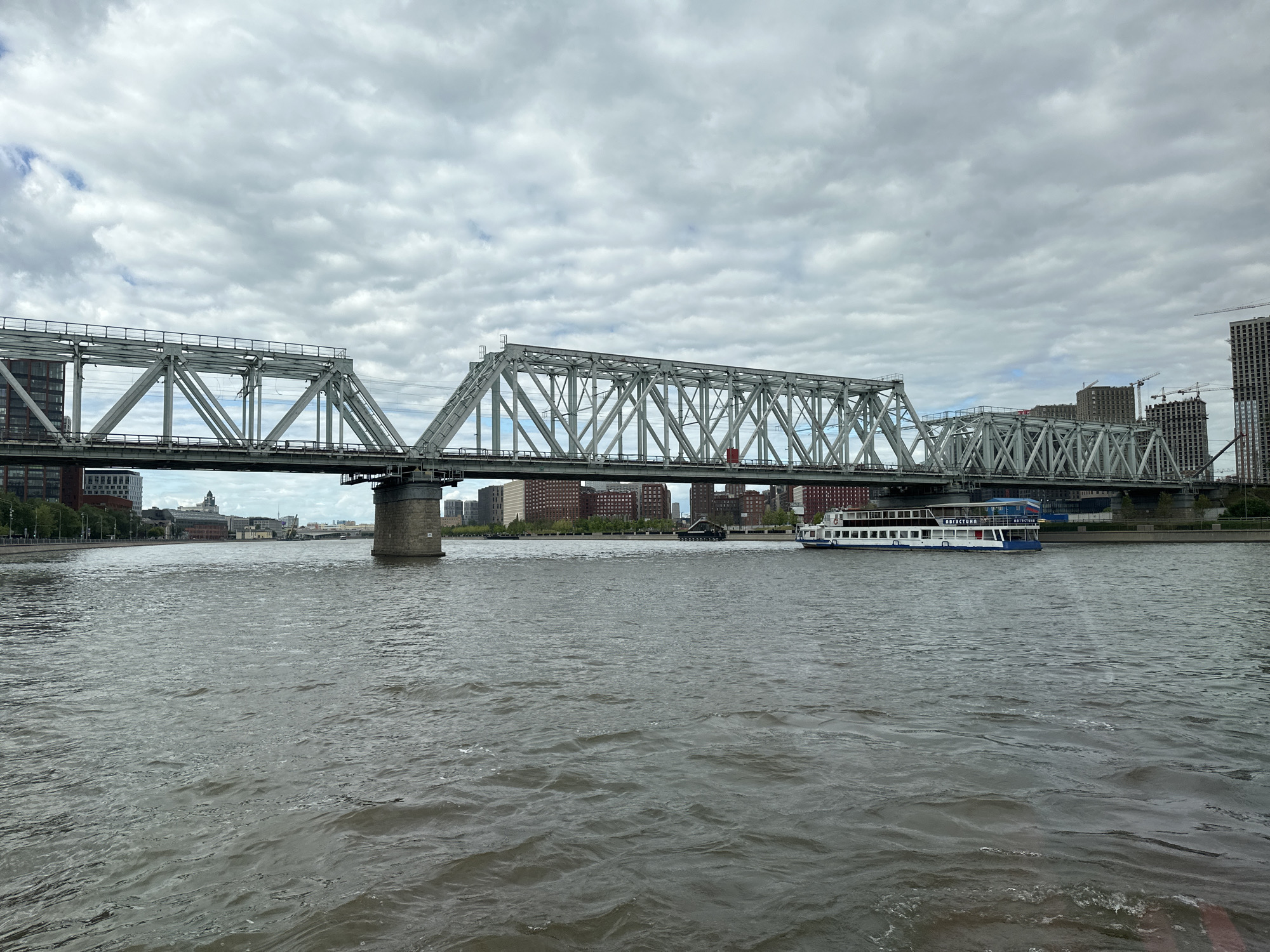 This media file depicts a wide view of a river with a large truss bridge spanning across it. The river is choppy, suggesting a breeze or recent activity. On the water, a white passenger boat with blue trim is visible, sailing towards the right of the frame. The boat has multiple decks and appears to be carrying passengers.
The bridge is a prominent structure, constructed with intricate metalwork, and railway tracks are visible on its deck. Beyond the bridge, a cityscape unfolds with buildings of various heights, including modern high-rises and older, more traditional structures. Construction cranes are visible among the taller buildings, indicating ongoing development. The sky is overcast with thick, grey clouds, suggesting a cloudy day. The overall scene is urban and industrial, with a focus on infrastructure and water transportation.
The text visible on the side of the boat reads "АВГУСТИНА" in blue Cyrillic letters. The location is Moscow, Russia, as indicated by the Cyrillic text and the architectural style of the buildings. The time of day is indeterminate due to the overcast sky, but the overall mood is neutral, focusing on the grandeur of the bridge and the activity on the river.