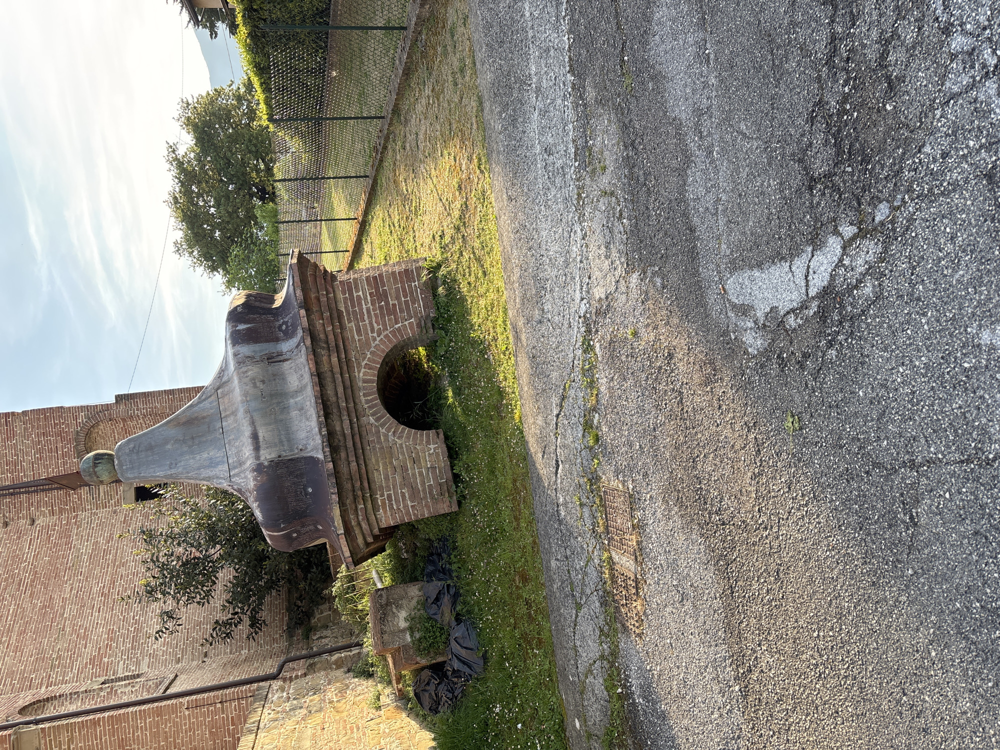This image, presented on its side, captures an outdoor scene, likely in Camerino, Italy, illuminated by the warm, golden light of either dawn or dusk. The sky is largely clear with a few wispy clouds. The central feature is a substantial, weathered metal structure, possibly a fountain or sculpture, shaped like an inverted bell or horn, topped with a spherical element. It is made of a dark, oxidized metal and stands on a brick base which includes a semi-circular opening. Behind this structure is a wall of aged red bricks, hinting at an historic building.
To the right of the brick base, an asphalt road stretches across the frame, showing signs of wear with visible cracks and patches. A narrow strip of grass, dotted with small white flowers and weeds, runs between the structure and the road. Several black plastic bags are strewn on this grassy area near the base of the brickwork. In the foreground of the road, a metal grate or manhole cover is partially visible. In the background, a large, vibrant green tree stands beyond a chain-link fence, under the expansive sky. No people or visible text are present, and the scene appears static.