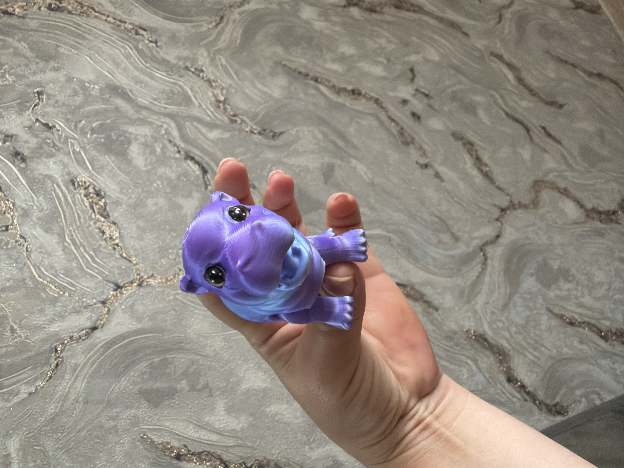 A person's hand is holding a small, purple, plastic toy hippo. The hippo is facing forward with its mouth open and its eyes are black. The background is a gray and white marbled wallpaper. The image is taken from a medium-high angle.