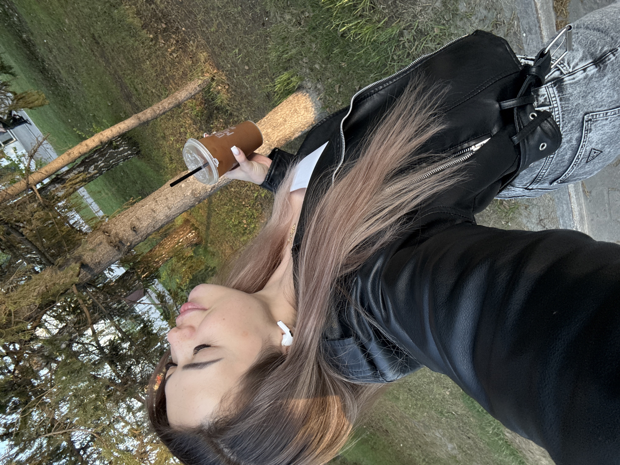 A woman stands outdoors, holding a coffee cup. She has long brown hair and is wearing a black leather jacket and gray jeans. She is looking to the side. The background includes trees and grass.