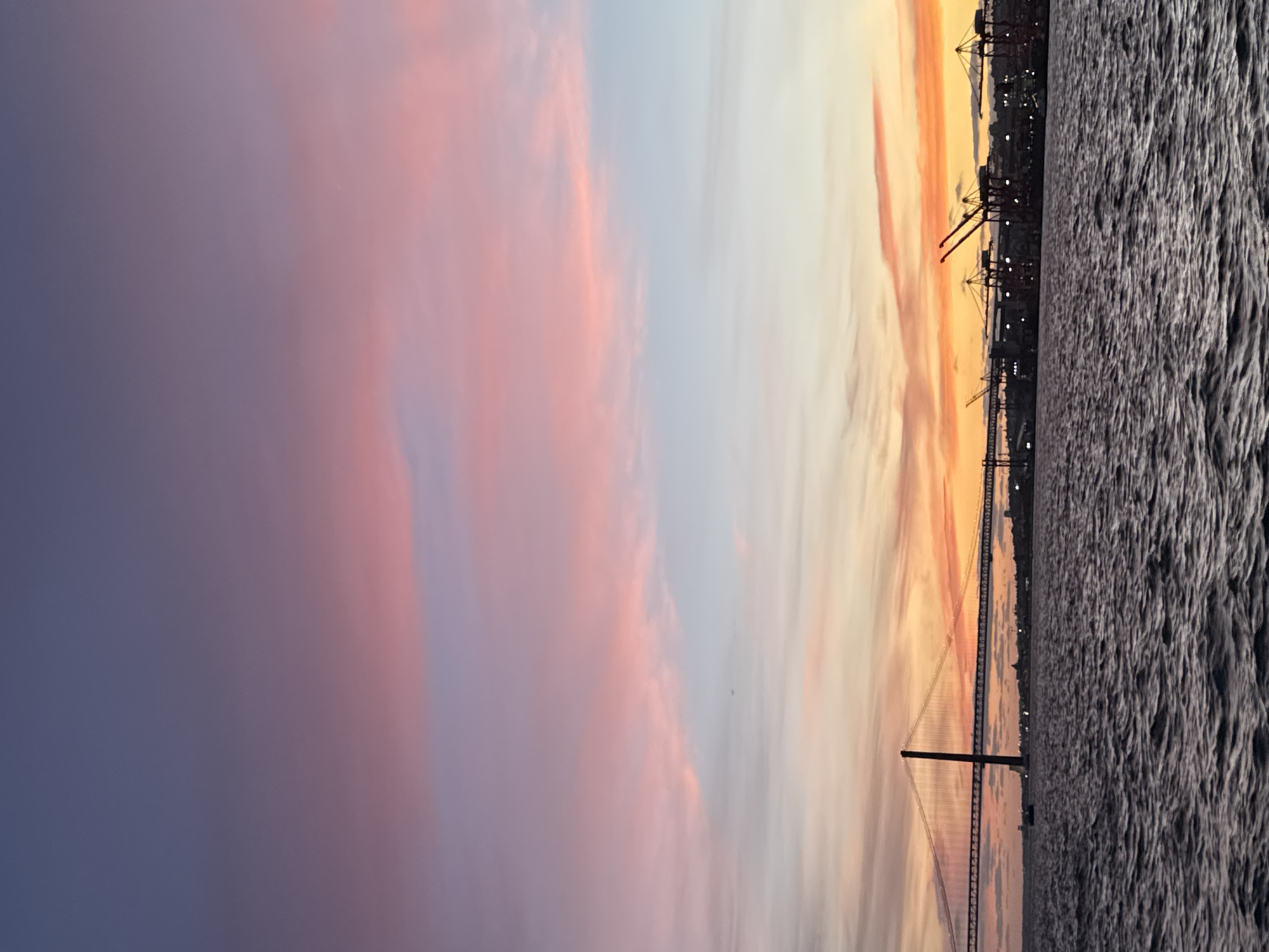 The image is of the 25 de Abril Bridge in Almada, Portugal, at sunset. The sky is a mix of pink, orange, and purple hues. The bridge is silhouetted against the colorful sky. There is water in the foreground, reflecting the colors of the sky. On the right, a dark cityscape is visible with cranes and buildings.