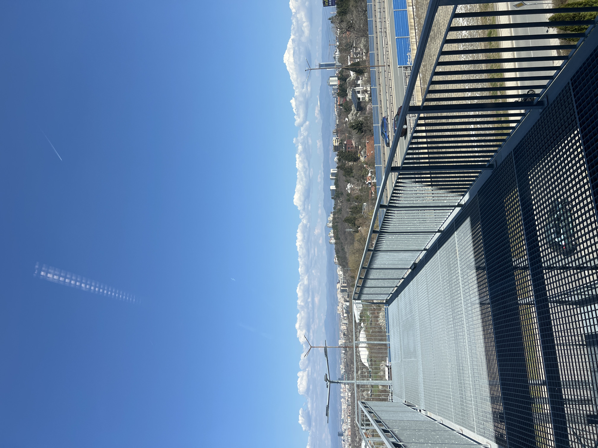 The image shows a view from a high vantage point in Sofia, Bulgaria, possibly a balcony or rooftop. A metal grid platform and railing are in the foreground, giving a sense of height and perspective. Below, there is a partial view of a road with cars and buildings interspersed with trees and green spaces. In the distance, there are clouds and a clear blue sky. There is a faint trail of a jet in the sky.