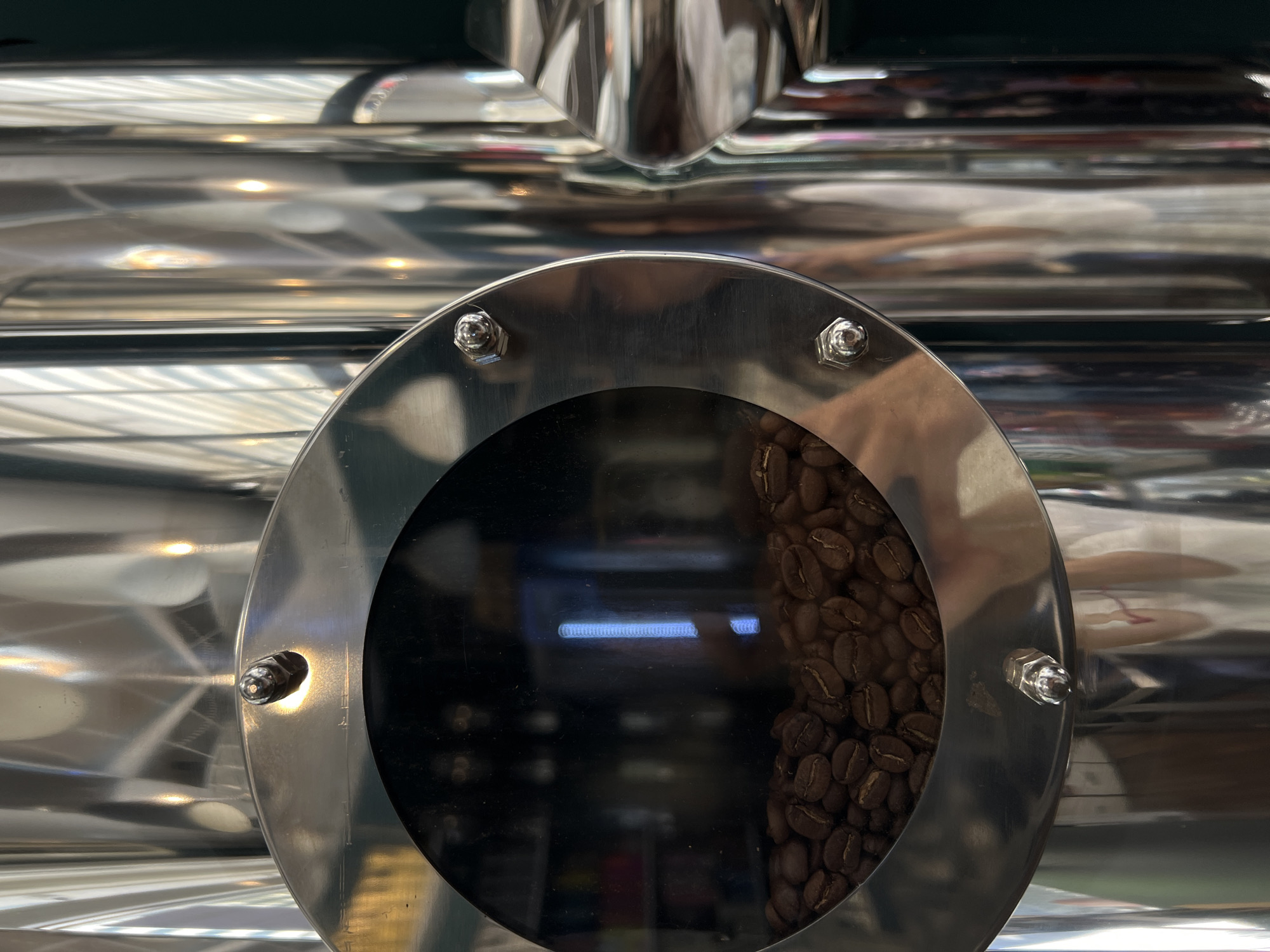 A close-up, vertical, eye-level shot features a metallic coffee roaster with a circular window showing roasted coffee beans. The window is secured with four visible bolts around its stainless steel rim. The background is dominated by the highly reflective, cylindrical body of the roaster, which mirrors light and vertical blinds. The scene is devoid of people and appears to be an interior shot within a commercial setting, potentially a coffee shop or roastery. The exact location in Sofia, Bulgaria, is not discernible from the visual information provided.
