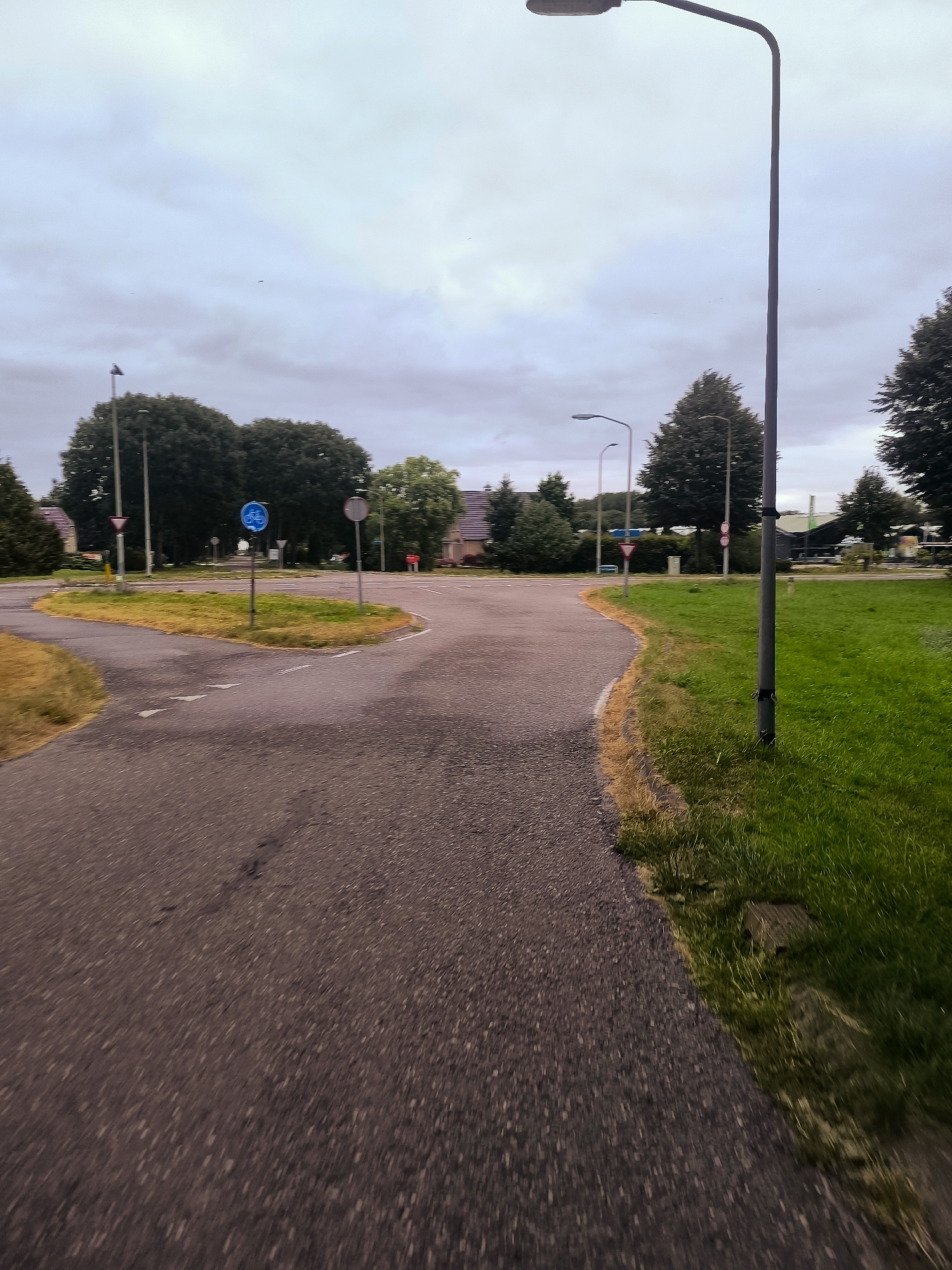 The image depicts a road intersection in Almere, Netherlands, likely in a residential or suburban area, under an overcast sky. A paved road curves from the foreground towards the left and right. To the right of the road is a well-maintained grassy area with a lamppost positioned near the edge. In the distance, several trees are visible, some with dense foliage, and there are residential buildings with tiled roofs. Traffic signs are present at the intersection, including a blue circular sign with a bicycle symbol and a red and white triangular yield sign. The overall atmosphere is subdued, suggesting it may be early morning or late afternoon, or simply a cloudy day. The perspective suggests the photograph was taken from a moving vehicle or while walking.