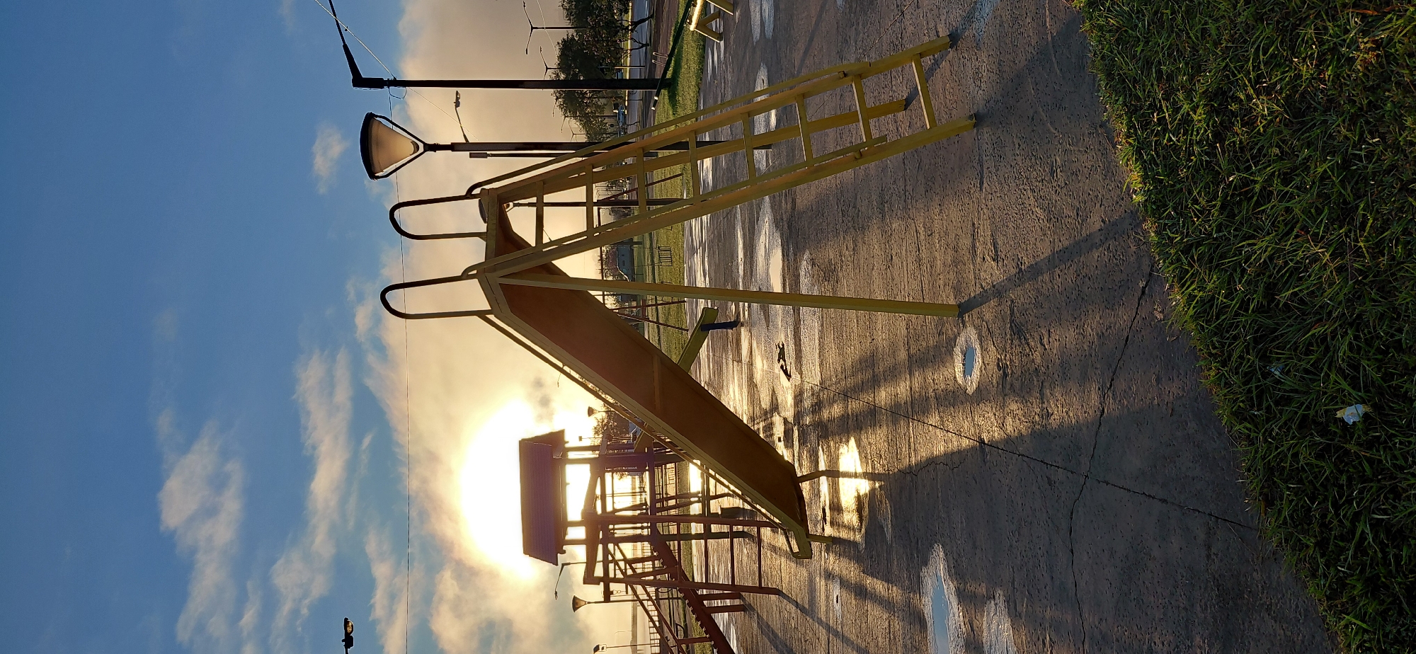 The image depicts a playground slide in the city of Encarnacion, Paraguay, at sunrise. The sun is low on the horizon, casting long shadows and creating a misty atmosphere over a body of water in the background. The slide, made of yellow metal, is positioned centrally in the frame, with its ladder leading up to the platform. Other playground equipment, including a wooden fort-like structure with a roof, is visible to the left. The ground is paved with concrete, showing patches of water and cracks. Streetlights are scattered throughout the area, one of which is illuminated, suggesting early morning or dusk. The sky is a clear blue with scattered white clouds. A thin strip of green grass is visible in the foreground. The overall scene is serene, with no people present.