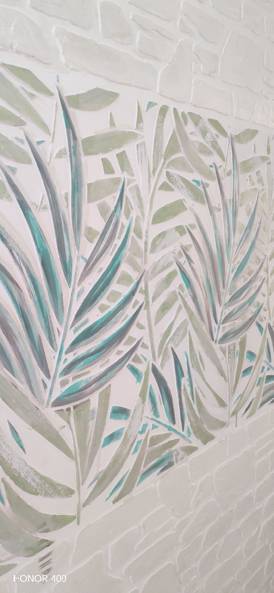 The image shows a wall with a textured, light-colored surface. The wall is decorated with a pattern of stylized leaves in shades of green and teal. The lower part of the wall has a brick-like texture. "HONOR 400" is visible in the bottom left corner.
There is not enough information to determine the location of this wall in Noviy, Russia. The image only shows the wall itself and does not provide any contextual clues about its surroundings or specific location.