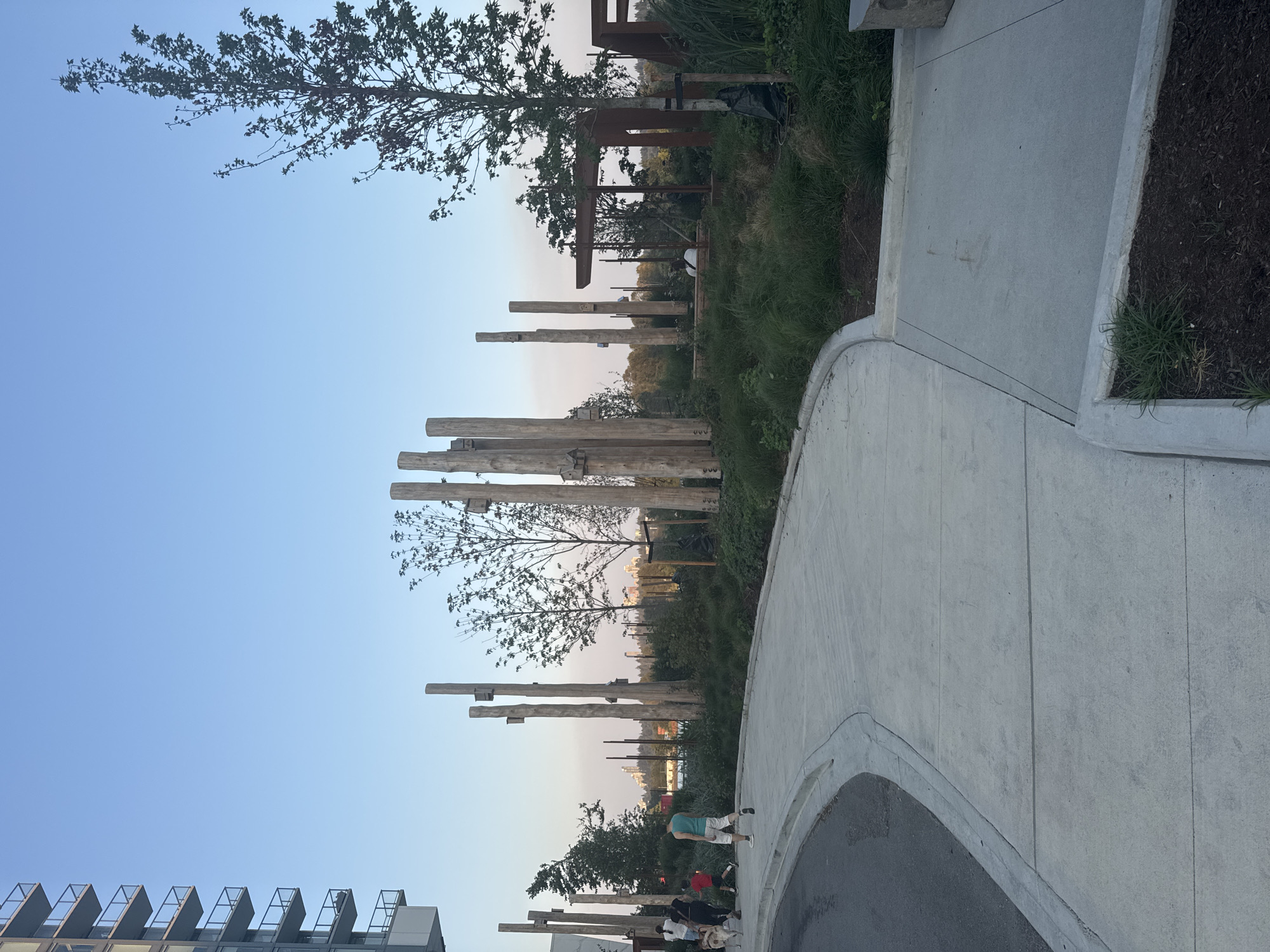 The image depicts a park scene in New Westminster, Canada, at dusk. A modern, multi-story building is visible on the left, with balconies and windows. In the foreground, a paved pathway curves to the left, bordered by a street on one side and grassy areas with strategically placed large wooden posts on the other. Several people are walking on the pathway. One person is walking a dog. In the background, a line of trees forms a natural boundary, and beyond that, city buildings are visible in the distance under a clear, pale blue sky. The park area features natural landscaping with tall grasses and some metal structures that appear to be part of the park's design, possibly for play or seating.