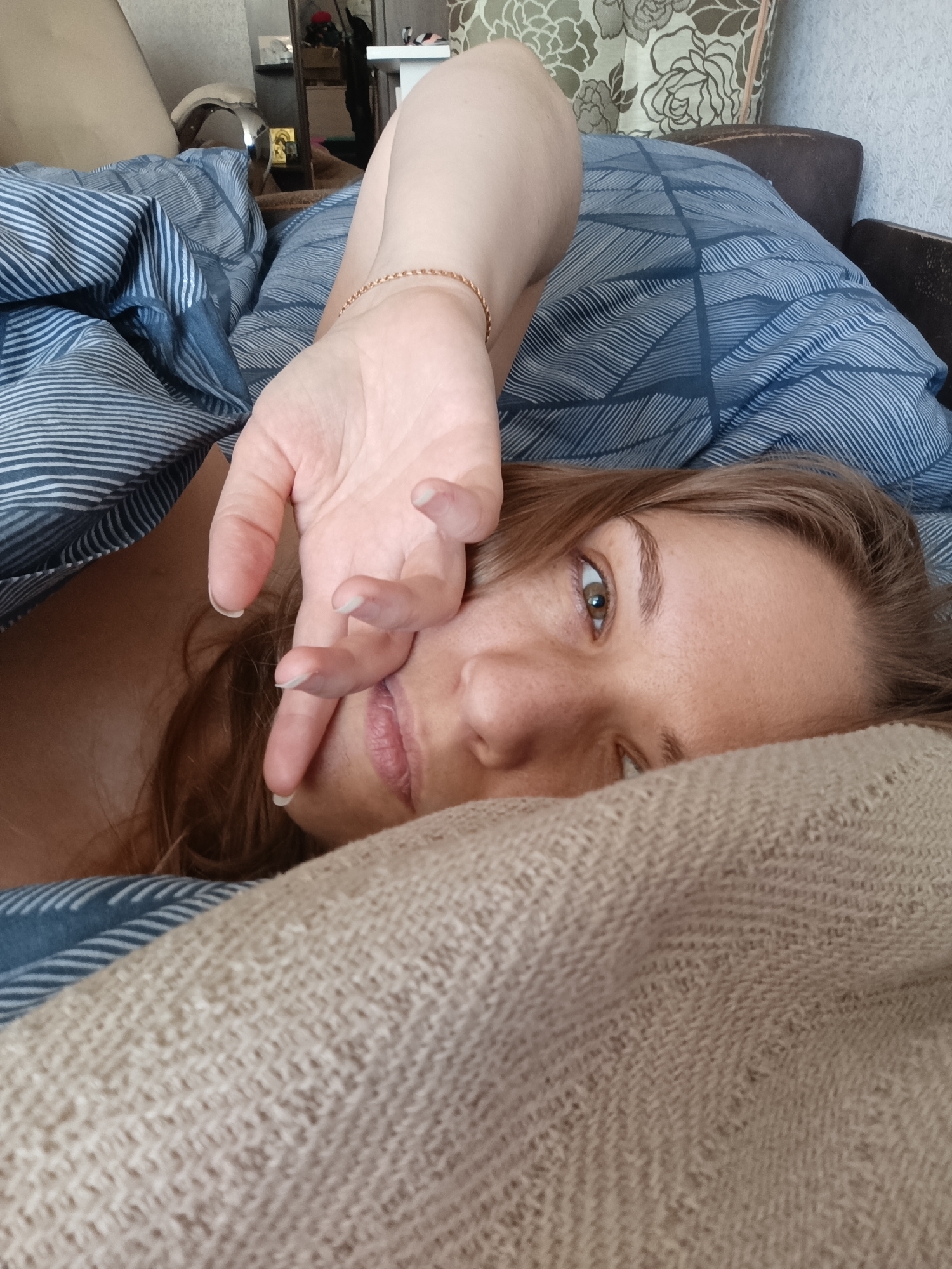 The image shows a close-up of a woman lying in bed. She is mostly covered by blue and white striped bedding and a beige knit blanket. Her right arm is raised, and her hand is partially covering her face, obscuring her mouth and nose. Her eyes are open and looking towards the camera. The lighting suggests it could be daytime. The background is blurred, but hints of furniture and floral-patterned curtains are visible, indicating an indoor setting, likely a bedroom. The woman's expression appears relaxed, perhaps indicating she is waking up or just lounging in bed. There is no visible text in the image.
