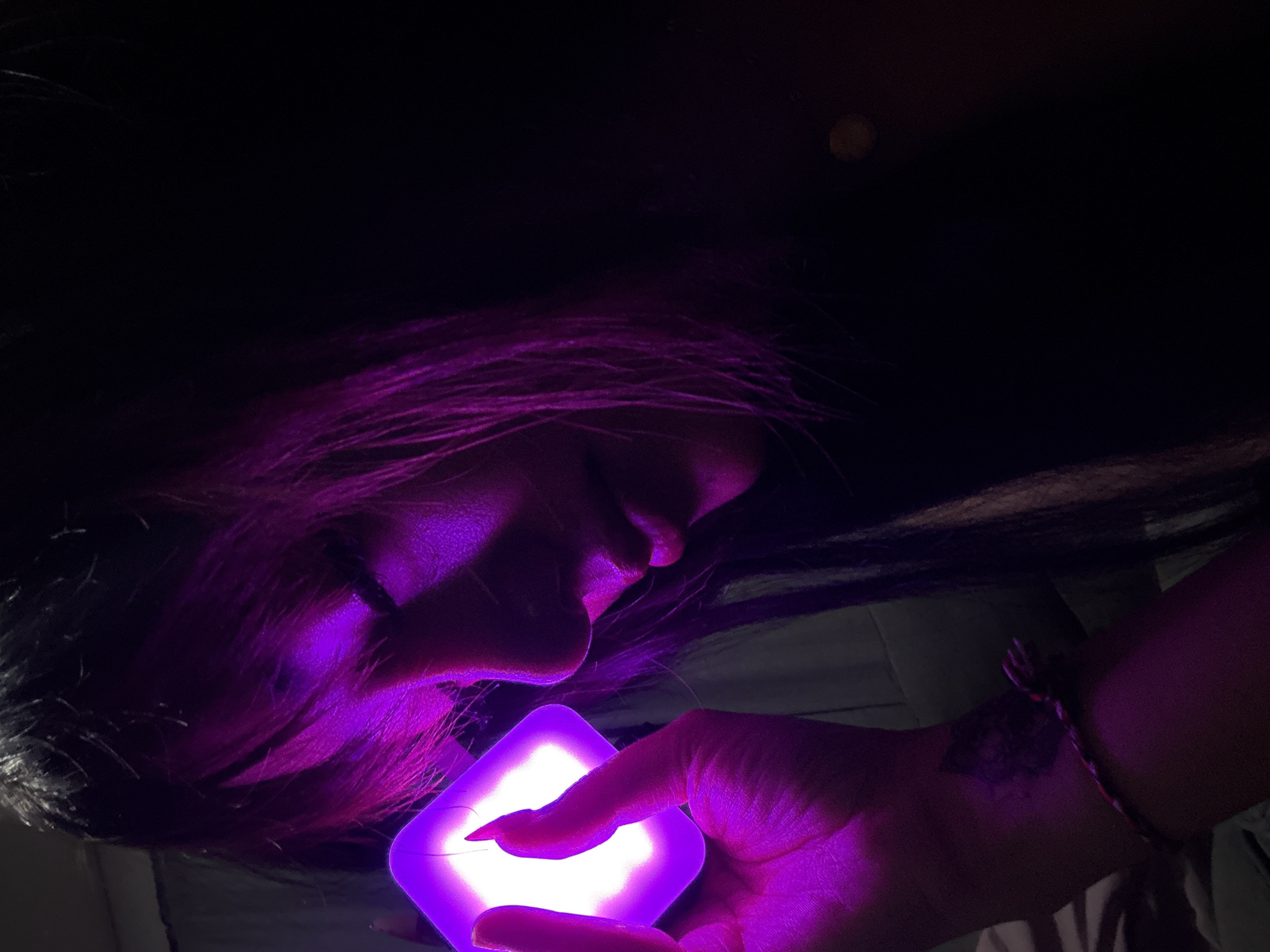 The image presents a close-up, intimate scene dominated by deep purple and magenta hues. A person, likely female, is shown in profile, their head positioned towards the left side of the frame and their body extending right. Their face is angled downwards, intently gazing at a glowing object held in their hand. The person's lips are slightly parted, conveying a sense of focus, wonder, or calm contemplation.
The individual's dark hair is illuminated by the light source, revealing striking magenta and purple tones, particularly around their head and shoulders. The background is largely dark, suggesting a nighttime setting or a dimly lit interior. A pale, possibly light-colored surface or fabric is subtly visible beneath the person's head and arm.
The central object is a square-shaped, intensely luminous device, emitting a strong purple light. It is held in the person's right hand, with one finger, featuring a pointed fingernail, gently touching its glowing surface. This primary light source casts a vibrant purple glow onto the person's face, hair, and hand, creating dramatic highlights and shadows.
On the person's right wrist, a braided bracelet can be seen, and further up their forearm, a dark, abstract-shaped tattoo is visible, partially illuminated by the purple light. The scene's dominant colors are shades of purple, magenta, and black. There is no visible text in the image. The overall mood is mysterious and centered around the captivating glow of the object.