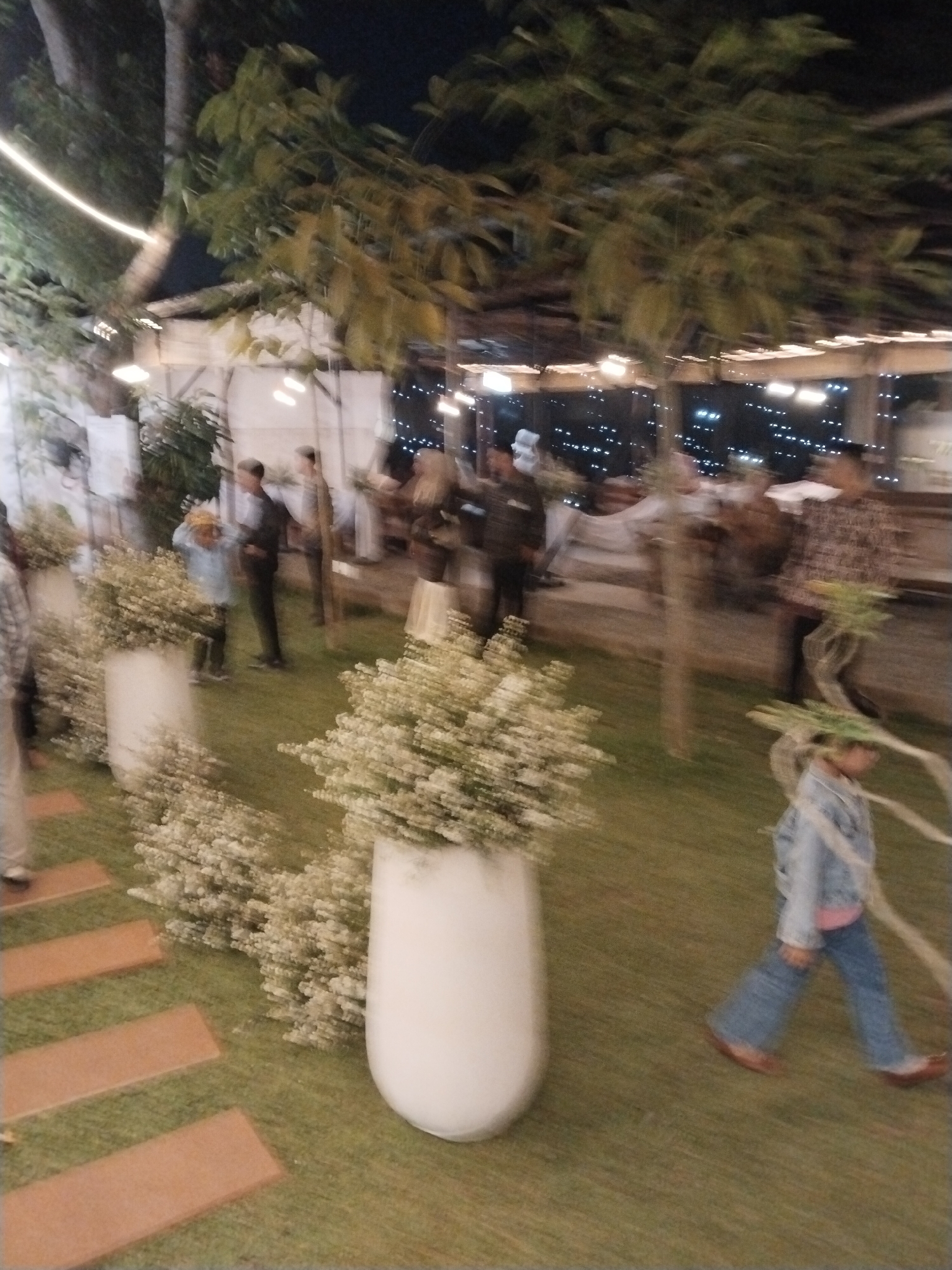 The image depicts an outdoor evening gathering in Serang, Indonesia. The scene is set on a grassy area with a stone pathway leading through it. Large white planters filled with white flowers are prominently placed, one to the left and one in the center foreground. In the background, several people are visible, some appearing to be in formal attire, mingling under trees with string lights illuminating the area. The lighting suggests it is dusk or night. A young child in a denim jacket and jeans is walking across the grass towards the right side of the frame. The overall impression is of a social event or reception held in a garden or outdoor venue.