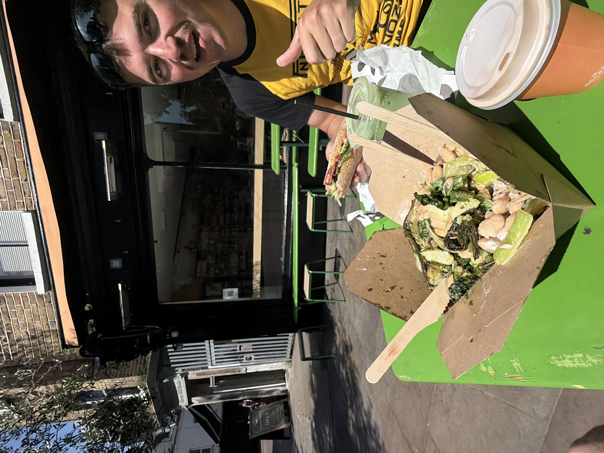 An eye-level, brightly lit outdoor photograph captures a person enjoying a meal at a green table. The central figure, a person with dark hair and sunglasses perched on their head, is smiling broadly at the camera and giving a thumbs-up with their right hand. They are wearing a yellow and black t-shirt, on which partial text like "NT" and possibly "LONDON" is visible.
On the green table in front of the person, there's an assortment of food and drinks. The person holds a half-eaten sandwich filled with greens and what appears to be meat. To their right, a clear cup with a black straw contains a green beverage, possibly a smoothie or iced tea. Further right, a brown takeaway box holds a salad-like meal with chickpeas, leafy greens, and other ingredients, along with a wooden fork. A paper coffee cup with a white lid, featuring the visible word "decent," is also on the table, next to a crumpled piece of white paper.
The setting appears to be an outdoor cafe or eatery. In the background, a dark storefront with large windows reflects the sky and surroundings. A brick building facade is visible on the left. Green metal chairs and tables are scattered on the paved area outside the building, indicating an outdoor dining space. Shadows suggest it is a sunny day, likely midday or early afternoon. In the lower background, a sign on the building clearly reads "OPEN," and below it, "Herne Hill Books" can be discerned, indicating a bookstore nearby in the Herne Hill area of London.
