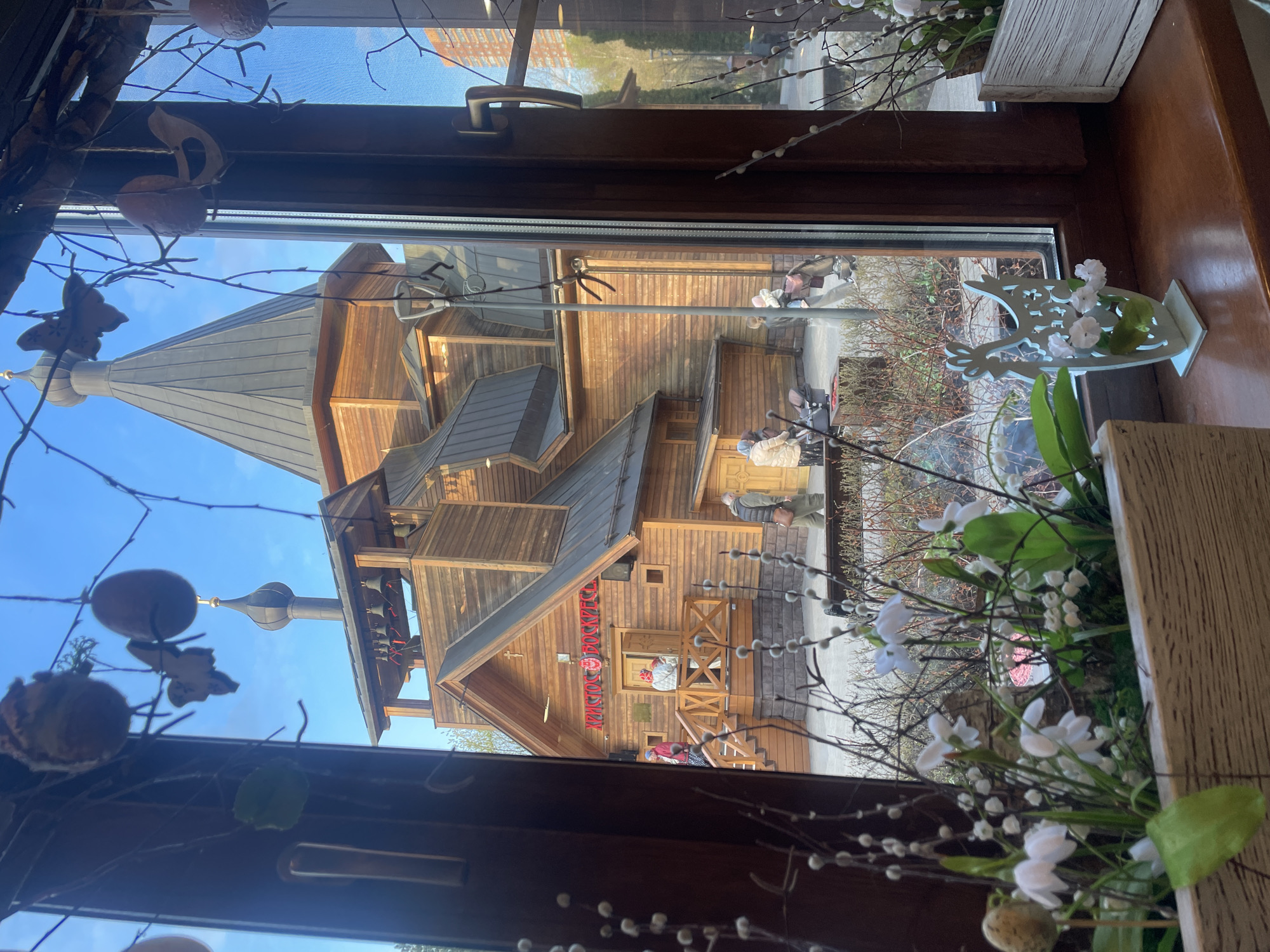 View through a window, showing the wooden Church of Peter and Fevronia, located in Khimki, Russia. The church has multiple tiers, with a grey roof and a tower. A few people are visible outside the church, near the entrance stairs. Inside the window frame, there are floral arrangements, including white flowers and branches. Some decorative eggs are also visible. The sky is blue.
