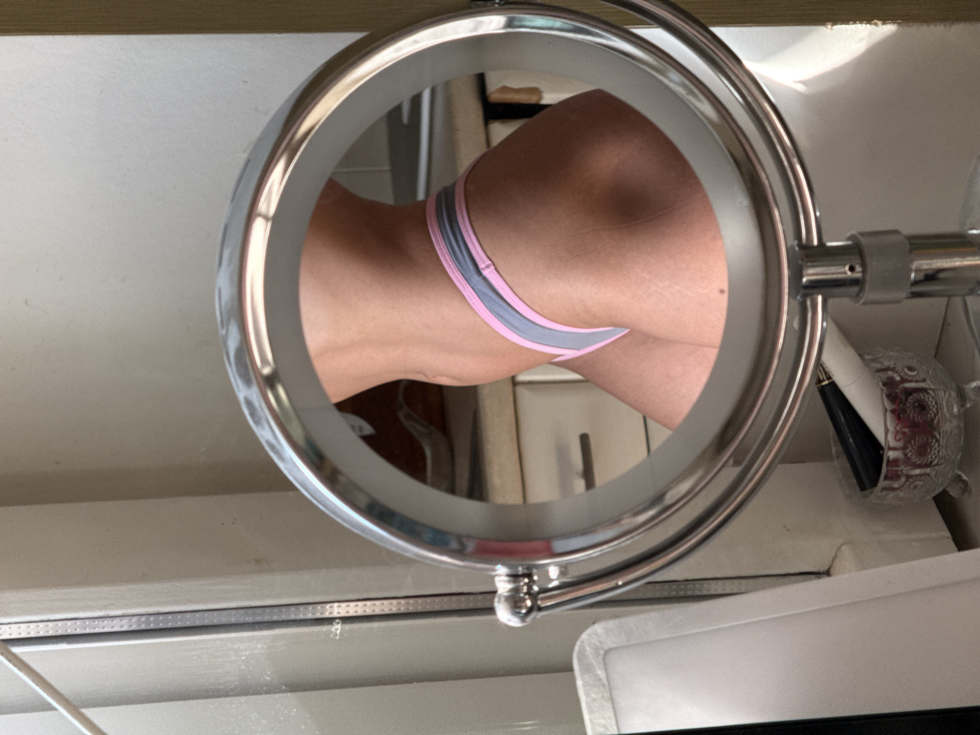 The image shows a reflection in a round, chrome vanity mirror. The reflection depicts a person's lower back and buttocks, clad in underwear with a gray and pink waistband. The background suggests a bathroom setting, with a cream-colored cabinet and wall. Additional items in the periphery of the mirror include a decorative glass holder with a dark object (possibly a makeup brush), and a white surface (possibly a sink or countertop). The location is not discernable from the information provided, and no information or implication of Saint Petersburg, Russia is present.