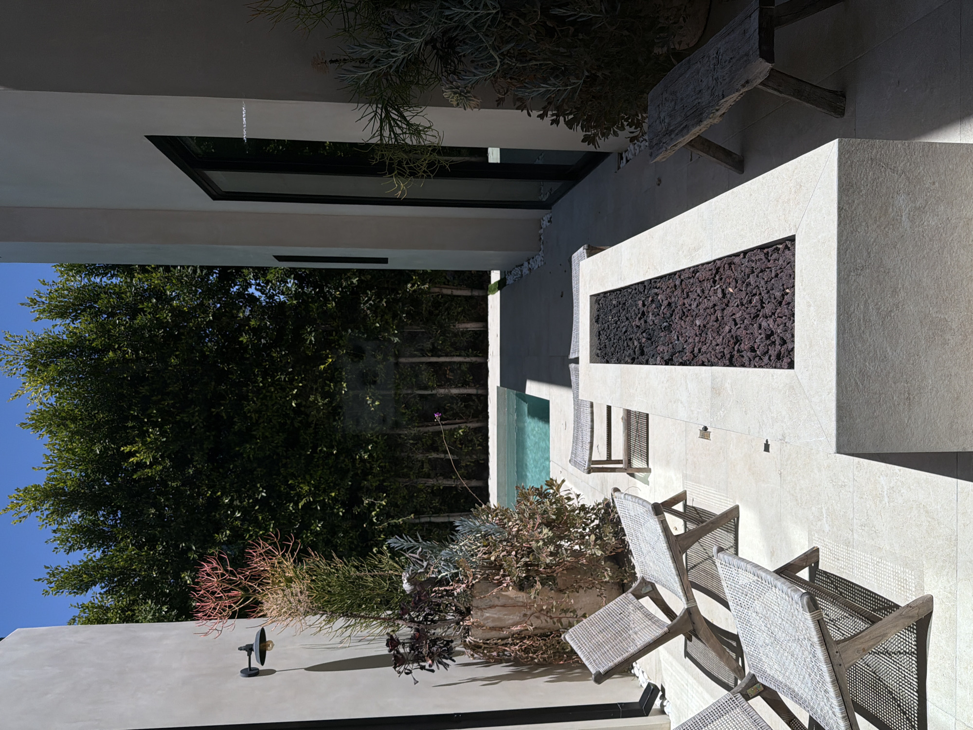 This outdoor scene, likely in Los Angeles, United States of America, features a modern architectural design with a fire pit as a central element. The fire pit is rectangular, constructed from light-colored stone or concrete, and filled with dark lava rock. Adjacent to the fire pit, several woven chairs are arranged, suggesting a seating area for relaxation or conversation.
The environment includes a swimming pool visible in the background, surrounded by lush greenery. Trees and plants create a sense of privacy and tranquility. The building's architecture is modern, with clean lines and a minimalist aesthetic. The presence of a light fixture on the wall and other architectural details indicates a well-designed and maintained outdoor space.