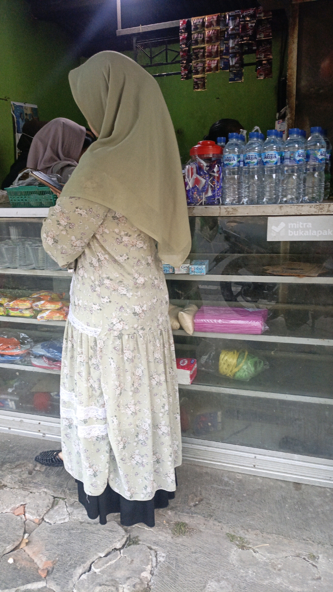 A woman wearing a light green hijab and a floral-patterned dress stands in front of a glass-fronted stall in Salatiga, Indonesia. The stall displays various products, including bottled water, snacks, and other packaged goods on shelves. A sign with the text "mitra bukalapak" is visible on one of the shelves. The ground in front of the stall is paved with cracked concrete and some patches of dirt and grass. The overall scene suggests a small local shop or roadside stall.