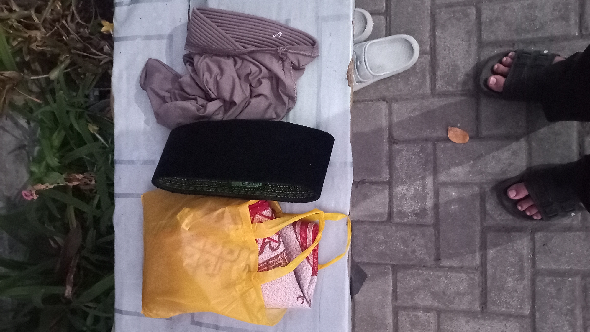 In the city of Pare, Indonesia, an overhead view captures a scene on a paved walkway, with lush greenery in the background. Laid out on a white cloth are several items: a yellow plastic bag containing folded fabric, a black peci (a brimless cap worn by Muslim men), and a folded brown hijab. Nearby, a pair of white sandals rests on the walkway. The foreground shows a pair of dark-colored sandals on the feet of an individual, with another person's feet also visible, suggesting a casual outdoor setting. A fallen brown leaf lies on the paving stones.