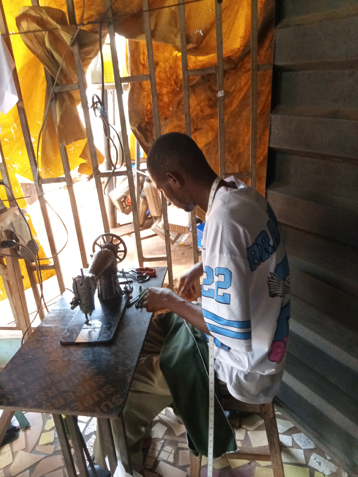 A man is sitting at a sewing machine in a workspace located in Nsugpe, Nigeria. The man is dark-skinned, with a shaved head, and is wearing a white jersey with a blue "22" and the word "BRAV" visible on the back. A white measuring tape is draped around his neck. He is seated on a simple wooden stool in front of a black sewing machine on a dark-colored wooden table with the word "SUPER" visible. Various tools, including a pair of pliers, are on the table near the sewing machine. Behind the man is a metal frame window covered with a yellow tarp. To the right, there is a gray metal staircase. The floor is covered with tile fragments arranged in a mosaic pattern. The setting appears to be a small tailoring shop.
