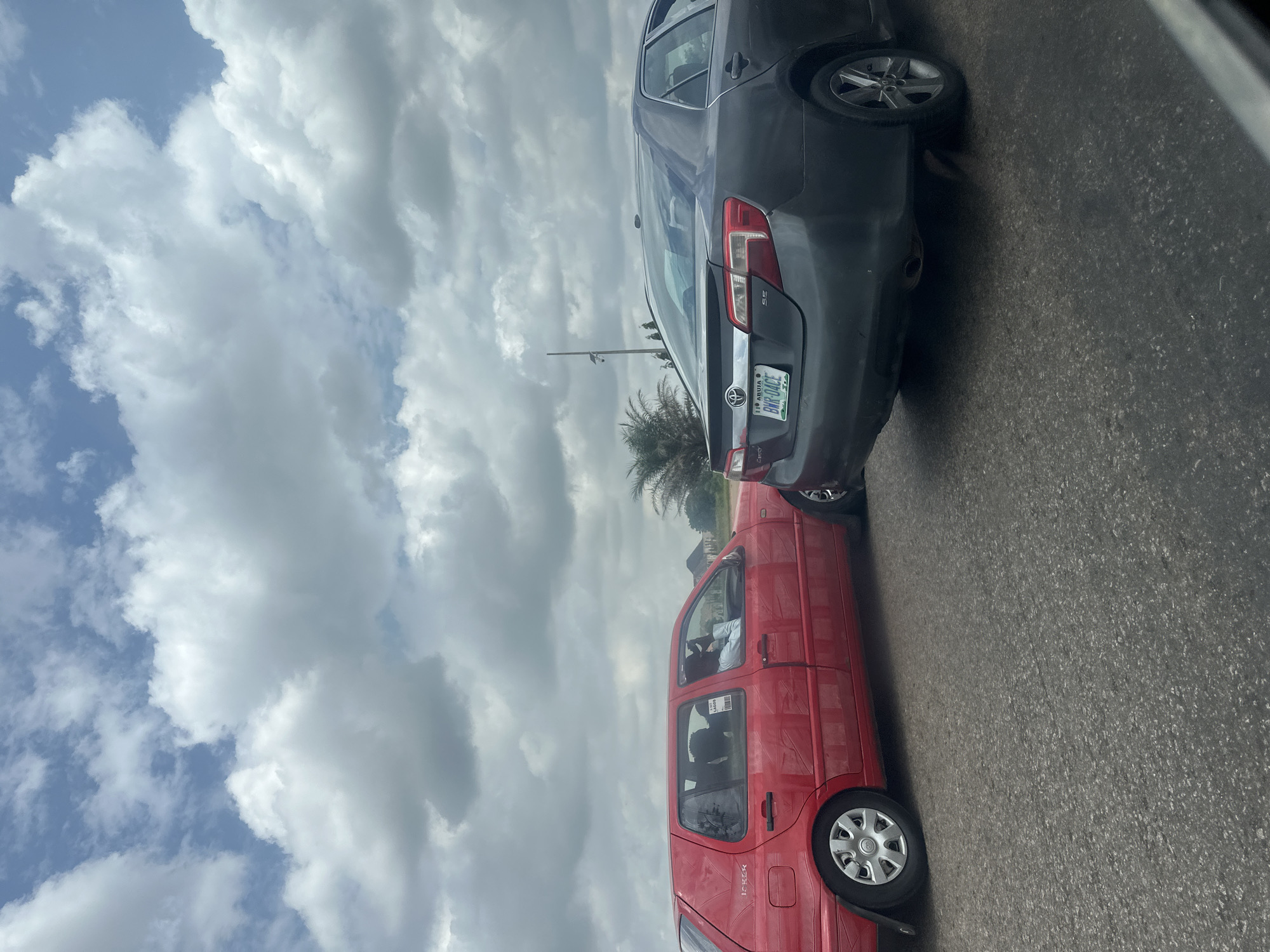 The image shows two cars on a paved road, likely in Abuja, Nigeria. The car on the left is a red Volkswagen Golf with a sticker on its rear side window. A person is visible inside the driver's seat. The car on the right is a dark gray Toyota Camry, seen from the rear. Its license plate reads "BWR 04CE" and indicates it is registered in Abuja. Palm trees and some buildings are visible in the background, under a cloudy sky. The road appears to be a multi-lane highway.