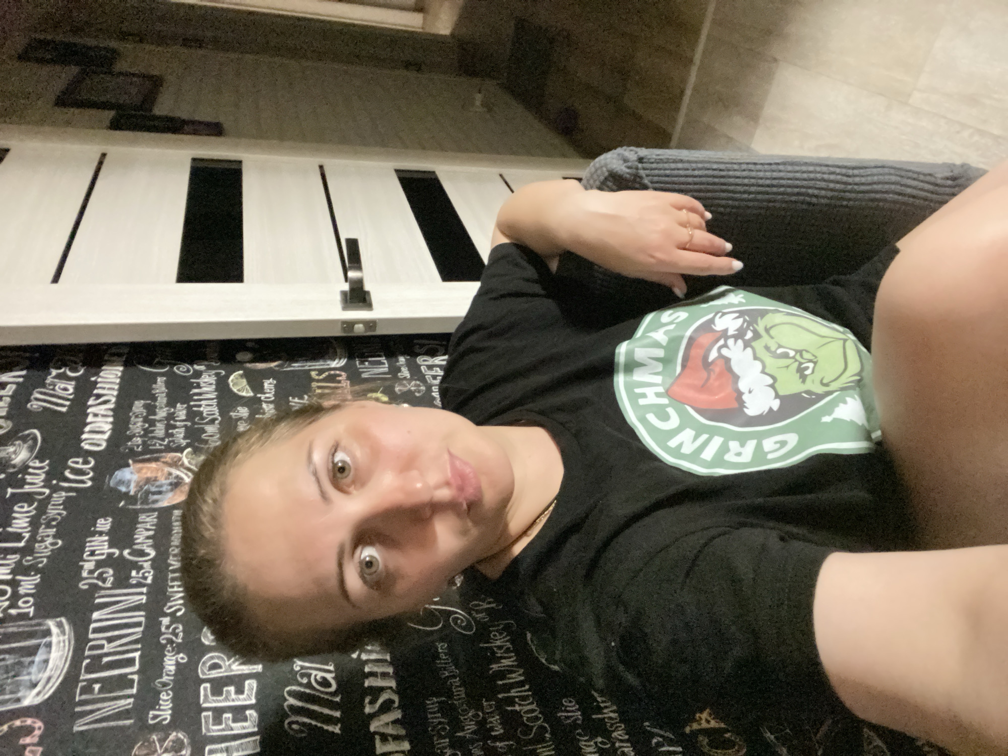A medium shot captures a woman in her 30s with light skin and light brown hair, seated in a dark gray armchair. She is wearing a black t-shirt with a green and white graphic of the Grinch wearing a Santa hat. The background features a dark wall with chalk art of cocktail recipes and text. To her right, a white interior door with horizontal black inserts is visible. The lighting is dim, suggesting an indoor evening setting. The image appears to have been taken in a residential interior.