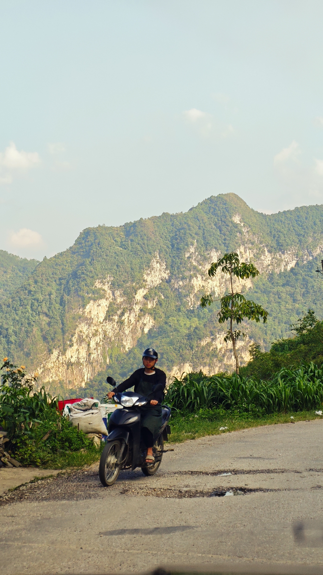 A man on a black scooter, wearing a black shirt, dark overalls, sandals, and a dark helmet, is on a road in Đường Thượng Commune, Vietnam. Behind him is a large mountain with visible white rock formations, partially covered with lush green vegetation. The road is cracked and shows signs of disrepair. Vegetation lines the sides of the road. The sky is a light blue with sparse clouds.