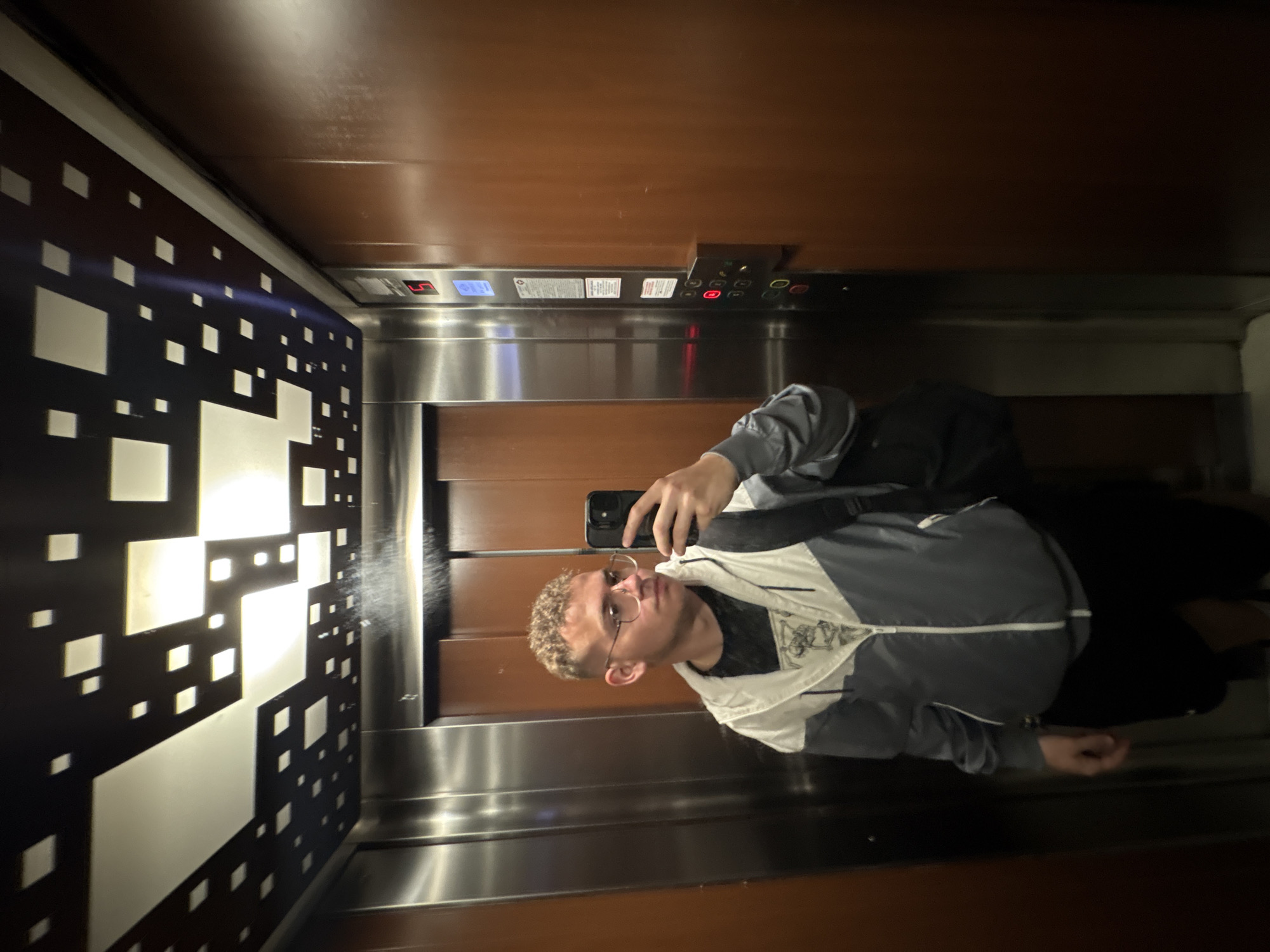 The image shows a selfie taken by a young person inside an elevator. The person, a male with short, light-colored curly hair and clear-rimmed glasses, is positioned slightly off-center and looks towards the camera. He is wearing a grey and white zippered jacket over a dark shirt, with a dark strap of what appears to be a backpack visible over his left shoulder. He holds a black smartphone in his right hand to take the picture.
The elevator's interior features a blend of textures and materials. The back wall and ceiling are clad in warm, reddish-brown wood paneling. The side walls and the panel containing the controls are made of brushed stainless steel, creating a somewhat reflective surface. On the left side wall, there is a distinct decorative panel, black in color, with various white illuminated squares forming an abstract pattern.
On the elevator control panel, a digital display prominently shows the letter "L" in red, indicating the current floor (likely the Lobby or ground floor). Beside it, a blue-lit display is also visible, though its exact content is indistinct. Below these indicators are a series of circular buttons, some of which are illuminated with red or green lights. The elevator is brightly lit, and the metallic surfaces reflect some of the interior light and the person. The scene depicts a casual moment in an elevator in Plovdiv, Bulgaria.