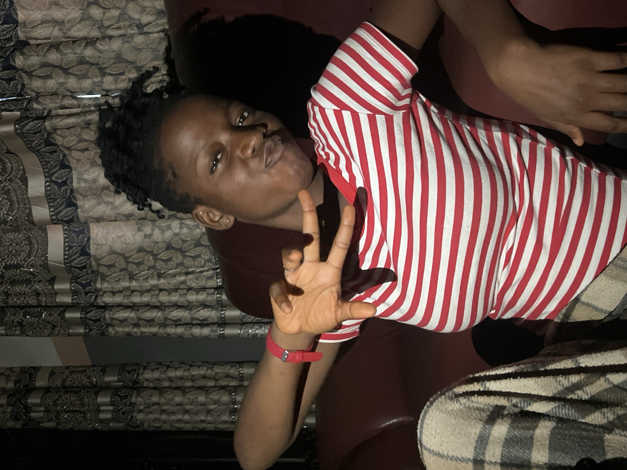 In Nkpor, Nigeria, a young Black woman with her hair styled in dark dreadlocks pulled up is captured in a medium shot, seated on a brown leather couch. She is wearing a red and white striped t-shirt. Her right arm is extended, and she is making a peace sign with her hand, which also has a ring on one finger. A red wristband is visible on her left wrist. A patterned blanket is draped over her left shoulder. Behind her, a decorative curtain with intricate patterns of beige and navy blue is visible. The lighting is dim, with highlights on her face and arms.