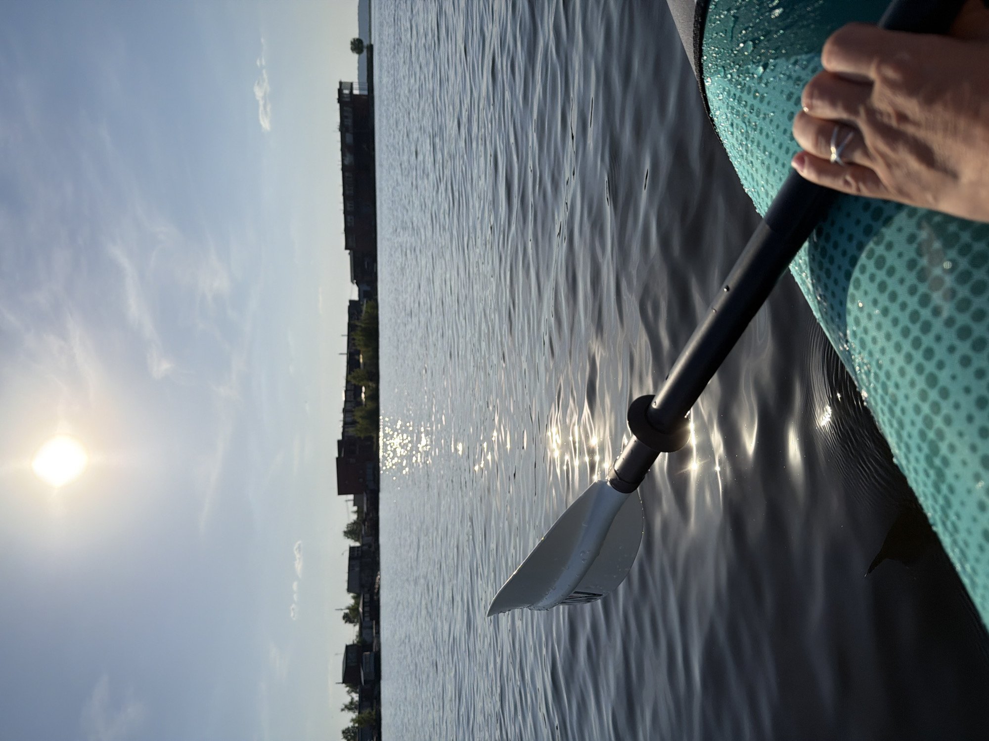 A slightly tilted, medium shot depicts a person paddling in a body of water. The sun shines brightly in the top left corner of the frame. The person's hand, with a ring on their ring finger, grips a black paddle shaft. The paddle blade is white and submerged in the water. A teal colored paddleboard is visible on the right edge of the frame. The water reflects sunlight. In the background, a row of buildings can be seen.
While it is difficult to determine the exact location of the image without additional information, the presence of buildings along the water's edge suggests a city location. The information provided suggests that this is the city of Chaikovsky, Russia.