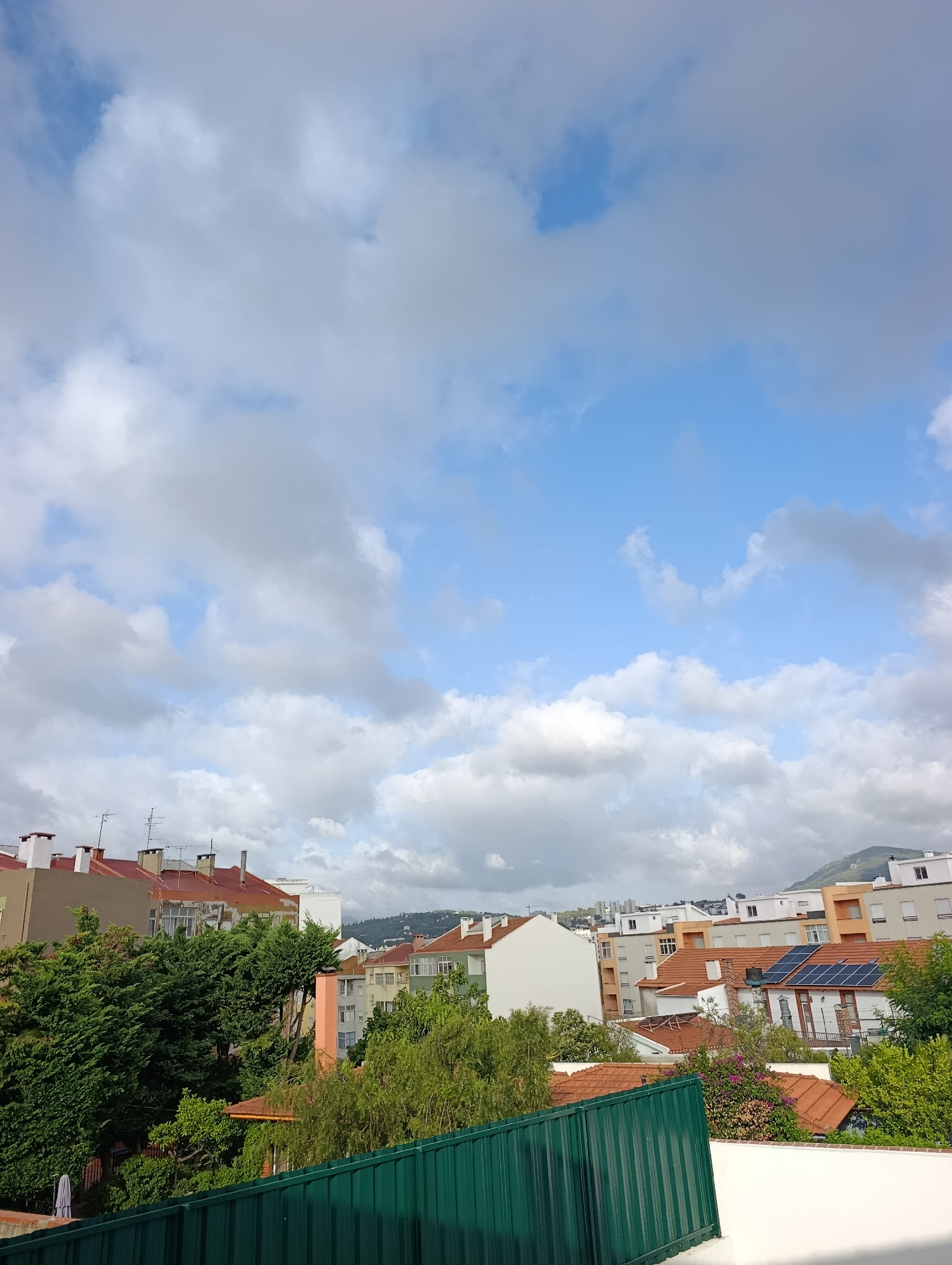 The image shows a view of a residential area with buildings and trees under a cloudy sky. In the foreground, a green metal fence runs horizontally across the bottom of the frame. Behind the fence, lush green trees are abundant, with some interspersed with flowering bushes. Beyond the trees, a collection of buildings with terracotta tiled roofs and various colored facades are visible. Some buildings have multiple stories, with windows and chimneys. A few solar panels are noticeable on one of the rooftops. In the background, a distant cityscape and a hilly landscape are discernible, shrouded in soft light from the overcast sky. The overall scene depicts a typical urban or suburban environment.