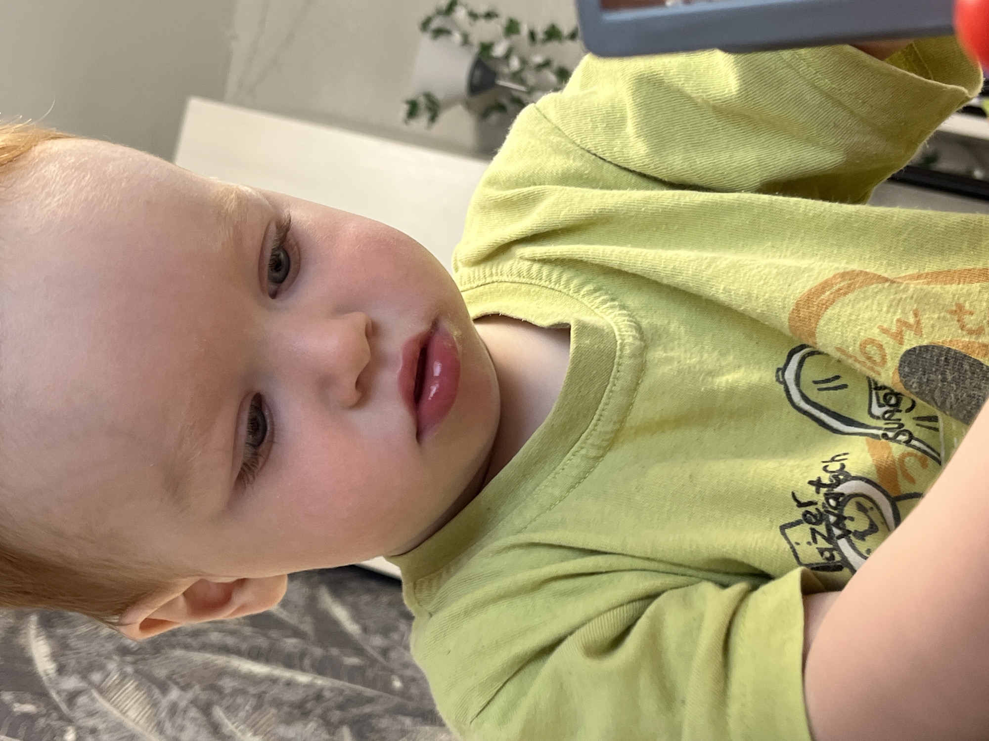 The image shows a close-up of a young child with fair skin and reddish-blonde hair, wearing a light green t-shirt with a graphic print. The child is looking down, with their head tilted slightly. Their lips are slightly parted, and there appears to be a light smattering of yellow around their mouth. The background is blurred, but suggests an indoor setting with a patterned curtain or tapestry on the left and a blurred white surface on the right, possibly a wall or furniture. There is also a hint of greenery in the background, potentially a houseplant. A gray object with a red circular element is visible on the right edge of the frame, possibly a toy or part of a larger object. The image is focused on the child's face and upper torso. There is no discernible information in the image to indicate a location in the city of Chaikovsky, Russia.