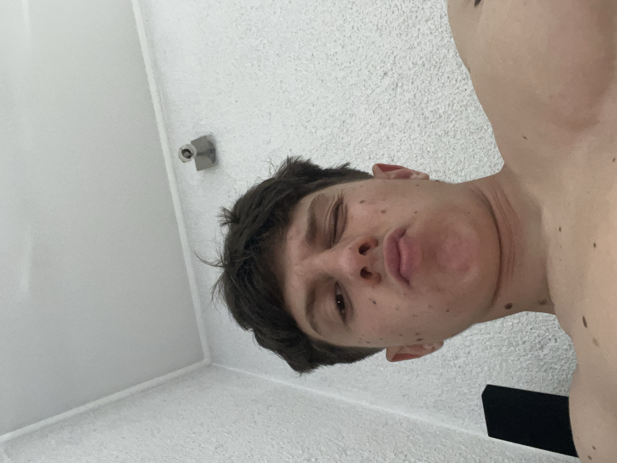 An overhead, low-angle photographic capture depicts an individual, presumed male, positioned indoors in a room located in Zlatograd, Bulgaria. The subject, viewed from the chest up, is shirtless, revealing visible shoulders and upper chest with multiple moles or freckles on the skin. The person has short, dark hair and is looking directly towards the camera, winking with the right eye and puckering the lips. The background consists of a light-colored, textured wall to the left and rear, meeting a smooth white ceiling with a defined corner molding. High on the rear wall, near the ceiling, a small, light-colored object with a dark base, resembling a sensor or small camera, is mounted. A partial view of a black, rectangular object, possibly an electronic device, is visible in the lower left portion of the frame.