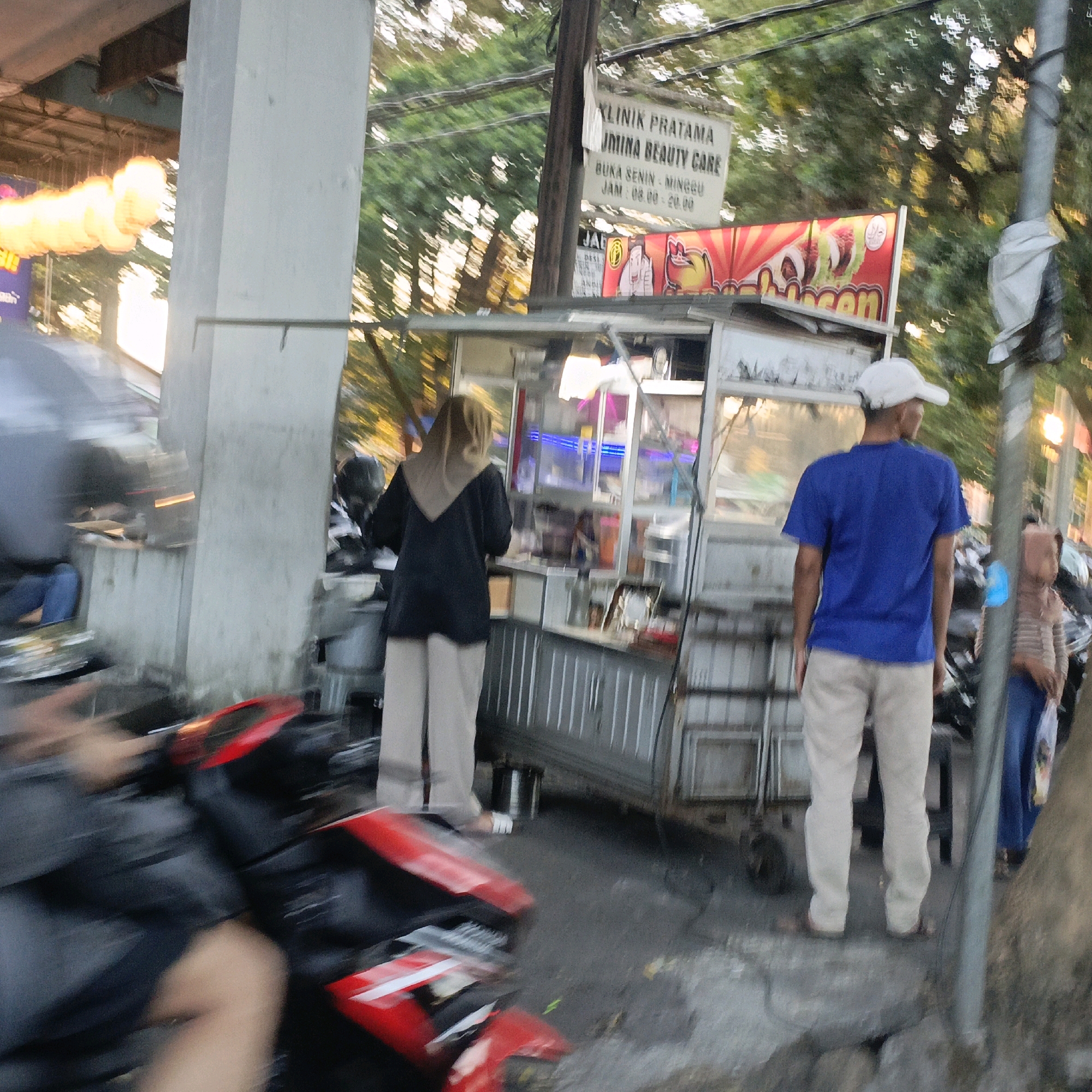 The image depicts a street scene in Salatiga, Indonesia. A street food vendor's cart is visible in the center, displaying food items and illuminated with neon lights. Several people are present: a woman wearing a dark top and light-colored pants is standing behind the cart, appearing to be the vendor. A man in a blue t-shirt and light-colored pants is standing to the right of the cart, facing away from the camera. Another person, partially obscured, is visible to the far right. In the foreground, a red motorcycle is parked, its front part slightly out of focus due to motion blur. A concrete pillar is prominent on the left side of the image, and trees and power lines are visible in the background, suggesting an urban environment. A sign in the background indicates a beauty clinic. The lighting suggests it is either early morning or late afternoon.