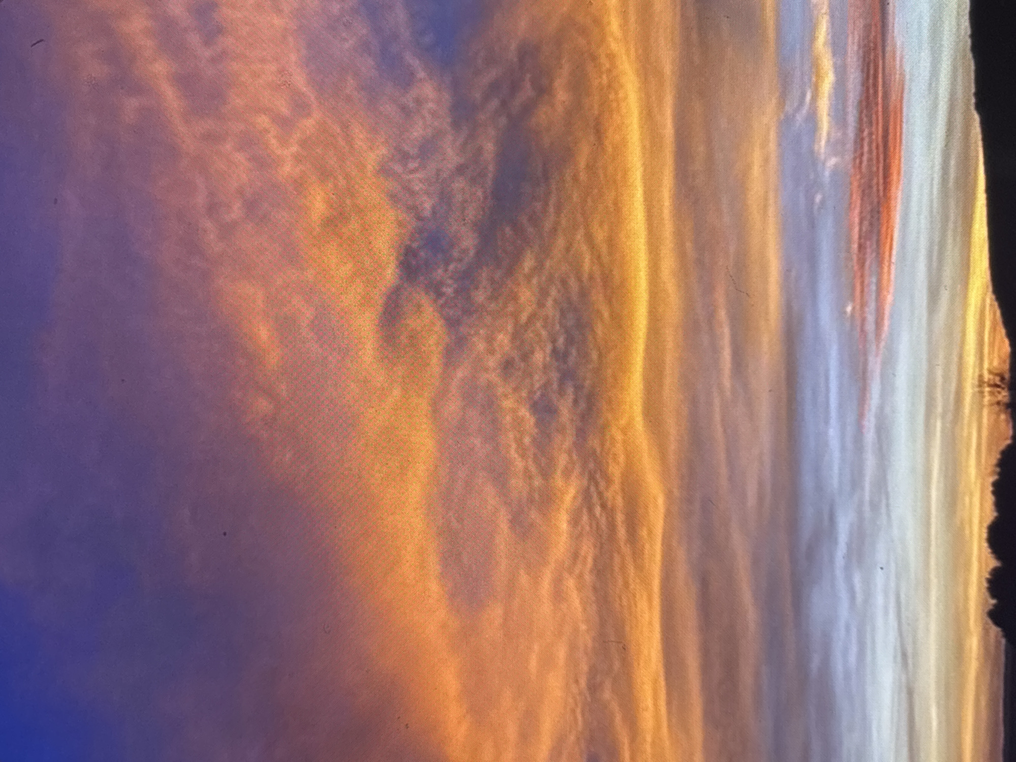 The image depicts a vibrant sunset, likely in Sofia, Bulgaria. The sky transitions from deep blue to orange, with pronounced cloud formations. The clouds are illuminated by the setting sun, creating a layered effect of orange and yellow hues. Some dark silhouetted foliage or trees are visible along the right edge of the image. The scene lacks people or clear objects, emphasizing the natural sky conditions.